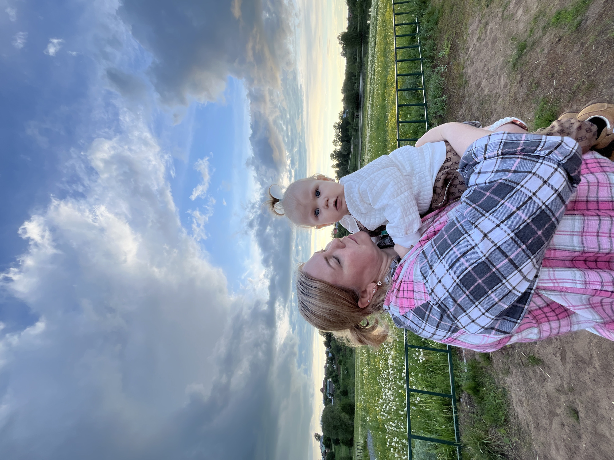 In Фоки, Russia, a woman holds a baby outdoors in a field. The woman has blond hair pulled back and is wearing a pink and gray plaid shirt. The baby has blond hair in pigtails. The background consists of a field of dandelions and a cloudy sky. A green metal fence is partially visible.