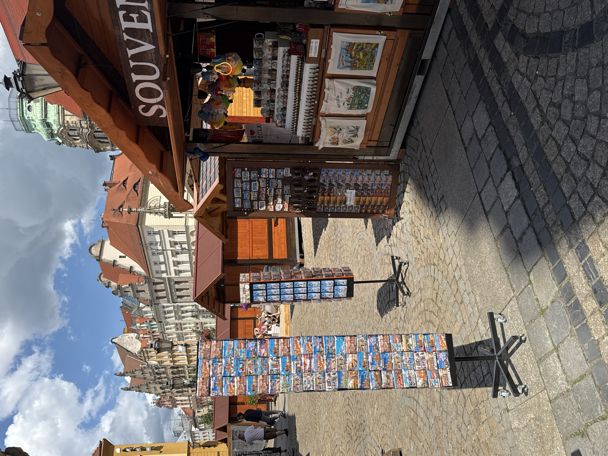 This image depicts an outdoor market scene on a cobblestone street in Wrocław, Poland, under a partly cloudy sky.
In the foreground to the right, a wooden souvenir stall with a brown roof displays various items. A sign partially reads "SOUVENIR". Inside the stall, colorful gnome figurines hang from the ceiling. Below them, shelves hold numerous small collectibles, including what appear to be miniature buildings, snow globes, and mugs. A price list is visible, indicating "1 FIGURINE - 20," "2 FIGURINES - 30," and "3 FIGURINES - 50." Three white canvas tote bags, each featuring a unique illustration of Wrocław landmarks and text such as "WROCŁAW POLSKA," are prominently displayed. To the left within this stall, a rotating display offers magnets and keychains, many with "Wrocław" themes.
In the midground, two black metal display stands with caster wheels are positioned on the cobblestones. The taller left stand is filled with postcards showcasing different scenes of Wrocław, with the city's name clearly visible on many. The shorter right stand displays smaller items, likely magnets or pins. Behind these stands, several brown wooden market stalls with reddish-brown roofs line the street; some are open, others appear closed. A person is seated at a table under an awning near one of these stalls in the background.
Further back on the left, two men are walking away from the camera on the cobblestone street; one wears a white t-shirt and shorts, the other a dark t-shirt and dark pants.
The background features a row of multi-story historic buildings with ornate facades and red-tiled roofs, characteristic of European market squares. A prominent green-domed tower, indicative of a historic building or church, rises above the stalls and buildings on the right side of the frame. The street is paved with grey cobblestones, some forming darker diagonal patterns, with small patches of grass growing between stones. Sunlight casts distinct shadows from the market stalls and display stands onto the pavement, indicating a bright day.
