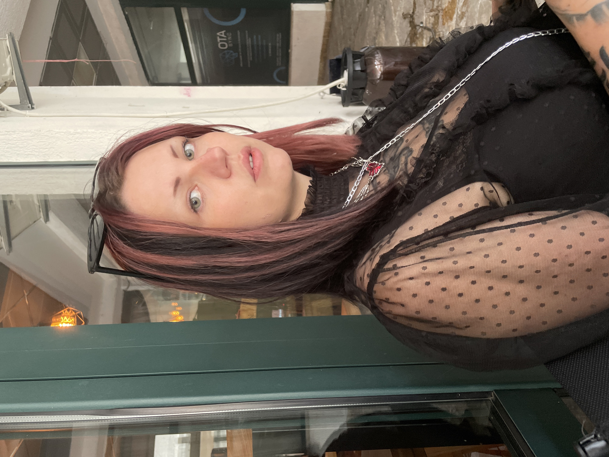 A woman with reddish-brown hair and fair skin poses for a photo. She is wearing a black dress with polka dot mesh sleeves and a silver necklace with a red gem pendant. Sunglasses are perched on her head. The background suggests an urban setting, possibly Budva, Montenegro. "OTA SYNC" is visible on a sign, which could be a business name. The overall scene is a casual outdoor shot.