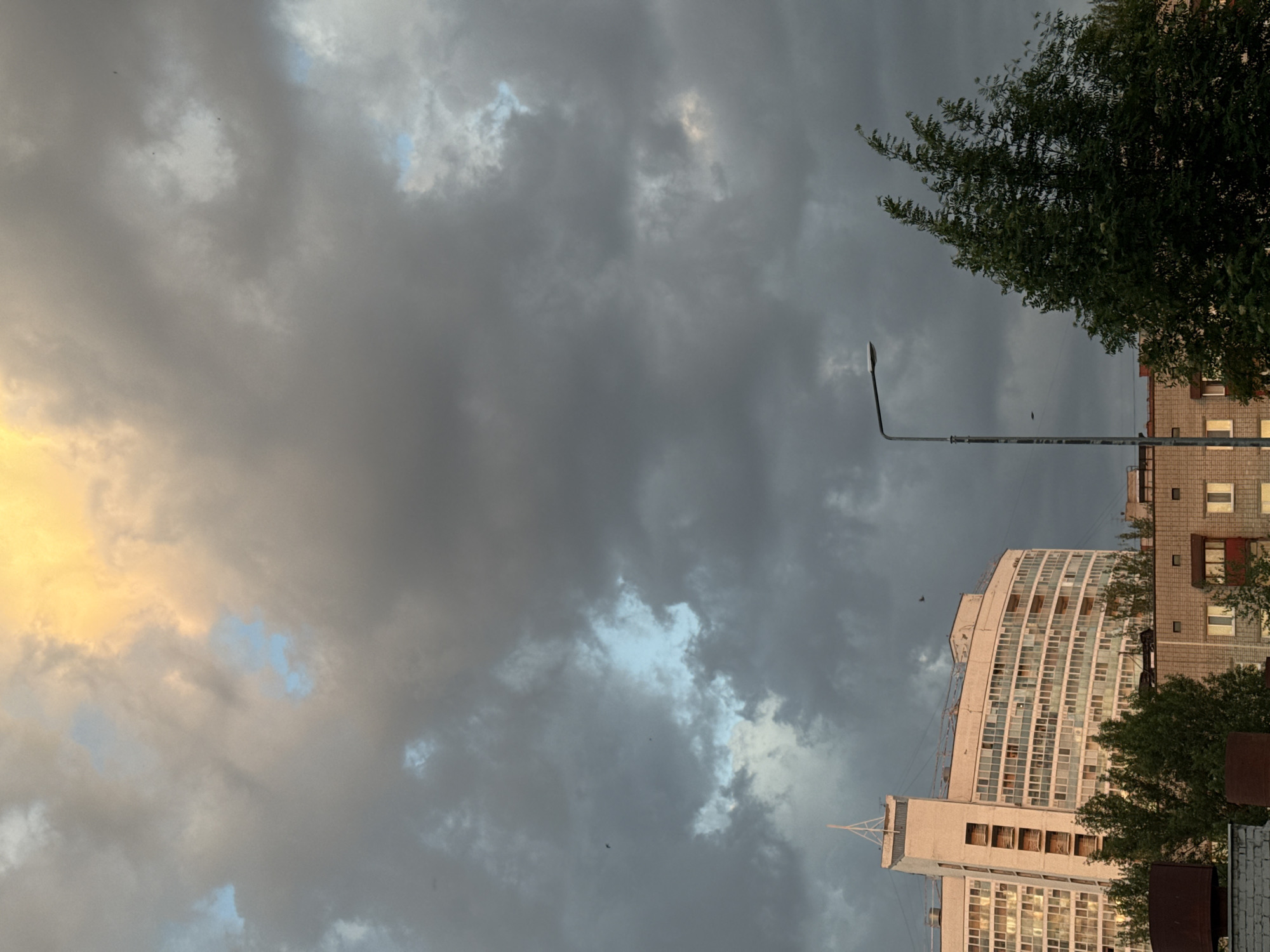 The image presents an upward view in a city environment, likely taken during a period of unsettled weather. The sky is dominated by a mix of dark, grey clouds, with small patches of lighter color and hints of blue sky visible through gaps in the cloud cover. To the right, portions of buildings can be seen, including what appears to be a multi-story apartment complex with numerous windows. The architecture style suggests a Soviet-era design common in many Russian cities. Adjacent to the buildings, the foliage of a tree is visible, adding a touch of greenery to the urban scene. A street lamp is also present, its metal pole cutting across the sky. Small birds are visible in the sky. The city could be Perm, Russia.