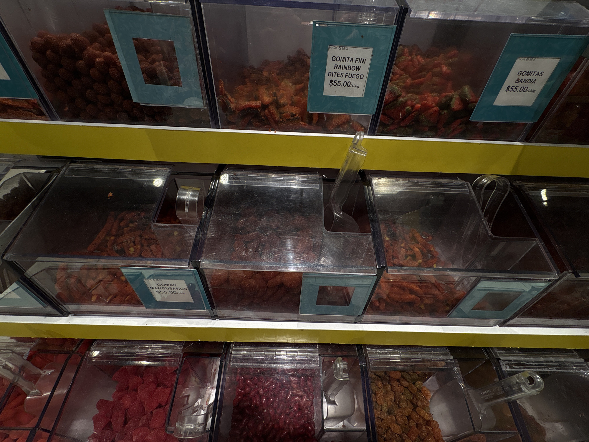 This close-up shot depicts a display of bulk candies, likely from a store specializing in confectionery in Mexico City, Mexico. The image is taken from a slightly elevated angle, showing two shelves of clear plastic bins containing various types of gummies and candies. Each bin has a sign indicating the name of the candy and the price per 100g (grams), priced at $55.00/100g, suggesting a premium or specialty product. The candies vary in color and shape. Some bins are nearly full, while others have been depleted, indicating customer interaction. Several clear plastic scoops are visible within the bins, indicating customers can self-serve the candies. The shelving units are a light yellow color, providing a backdrop for the colorful display of candies.