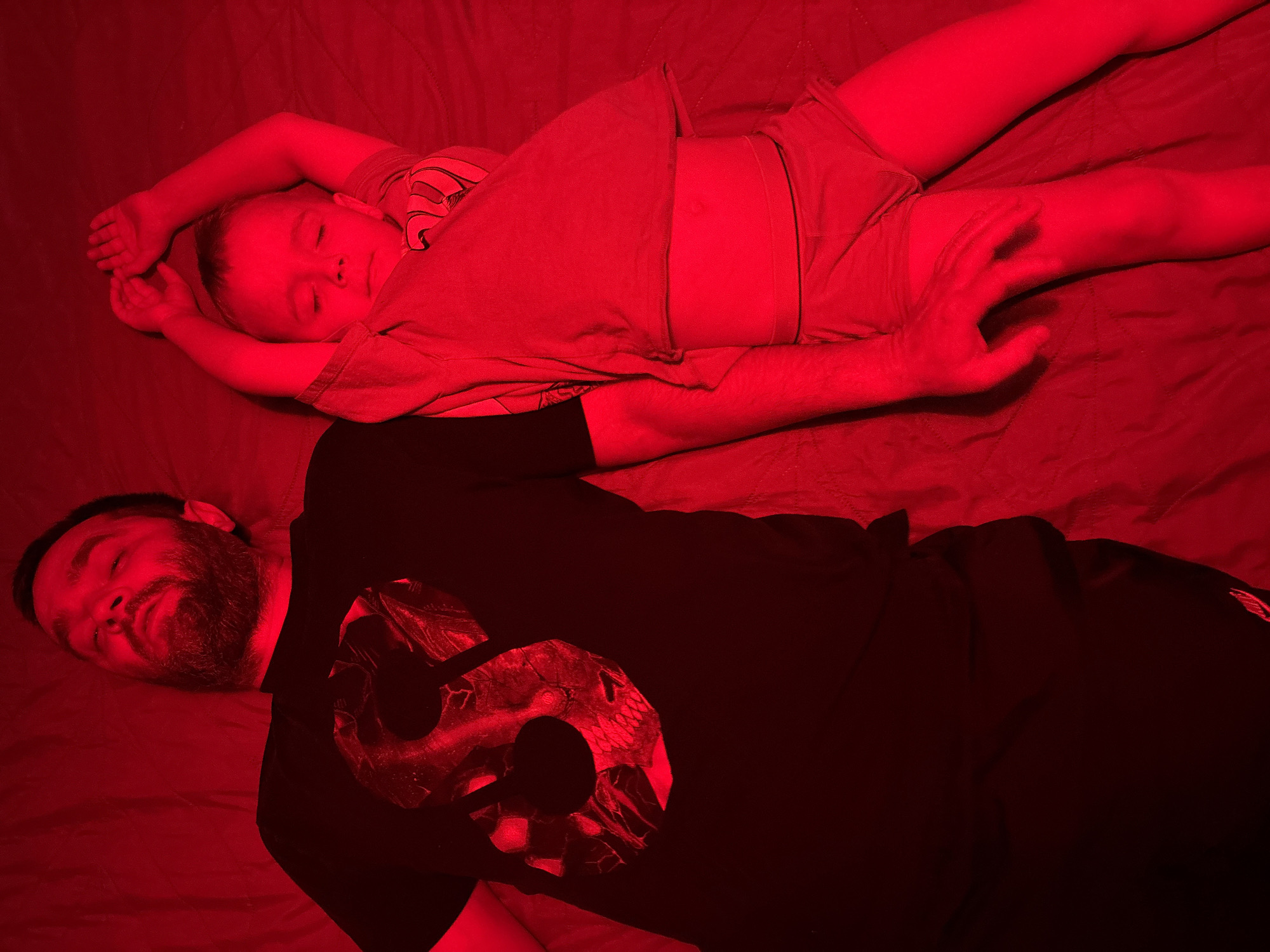 A man and a child are lying down next to each other on a bed. Both appear to be sleeping. The man is wearing a black t-shirt with a large, circular design on the front and black pants. He has a beard. The child is wearing light-colored shorts and a t-shirt. The lighting in the room is predominantly red. There is no indication of the location being in Chaikovsky, Russia.