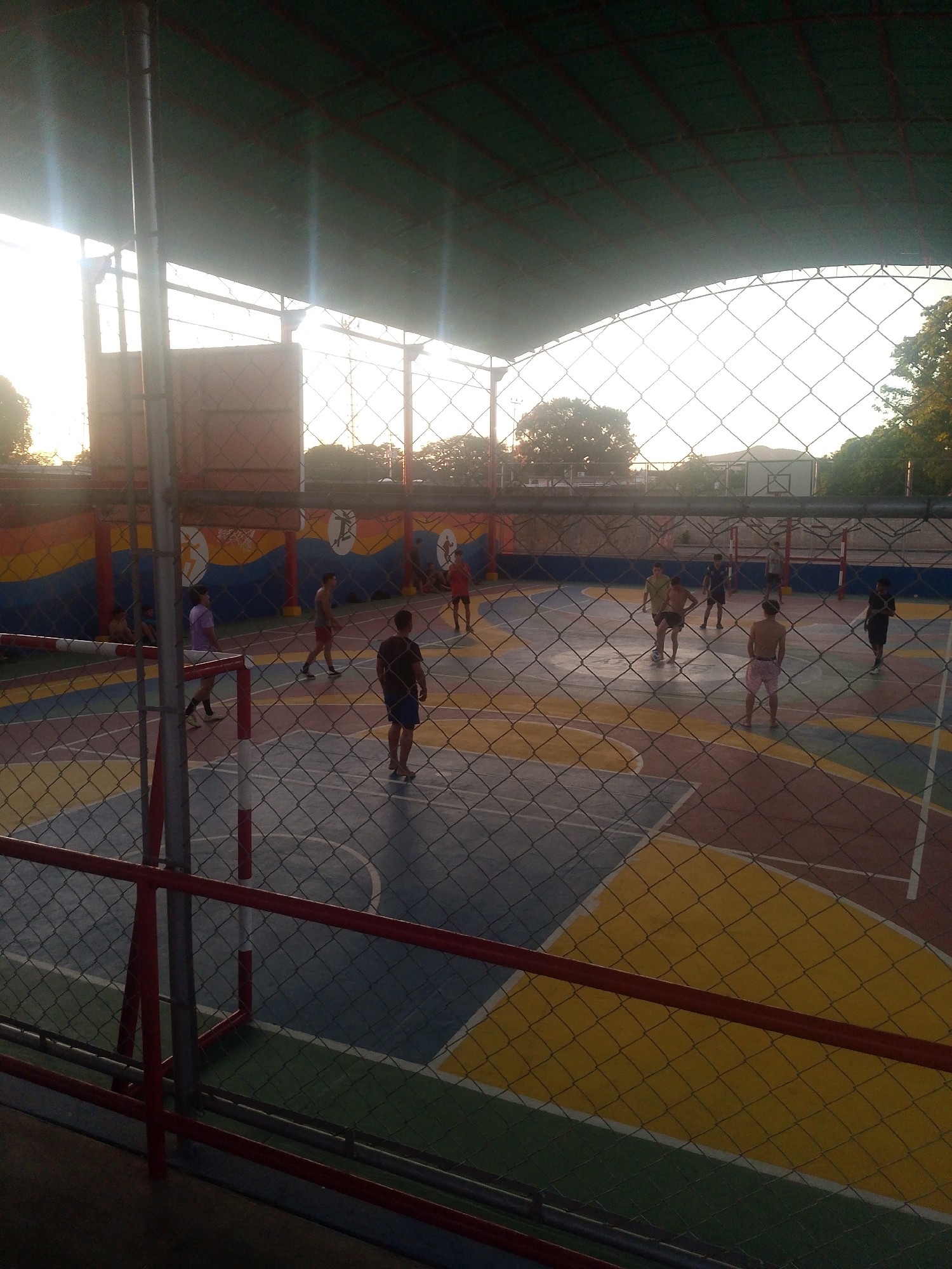 The image shows a group of people playing on a multi-colored court enclosed by a chain-link fence, likely in Juan Griego, Venezuela, based on the city attribution. The court is under a covered structure. There are approximately ten individuals visible in the playing area, some appear to be actively participating in a game. The court has a basketball hoop on the left side. The walls surrounding the court are painted with colorful murals. There is a building or structure visible behind the fence in the background. The light suggests that the image was taken during the day, possibly early evening. A red railing is in the foreground.