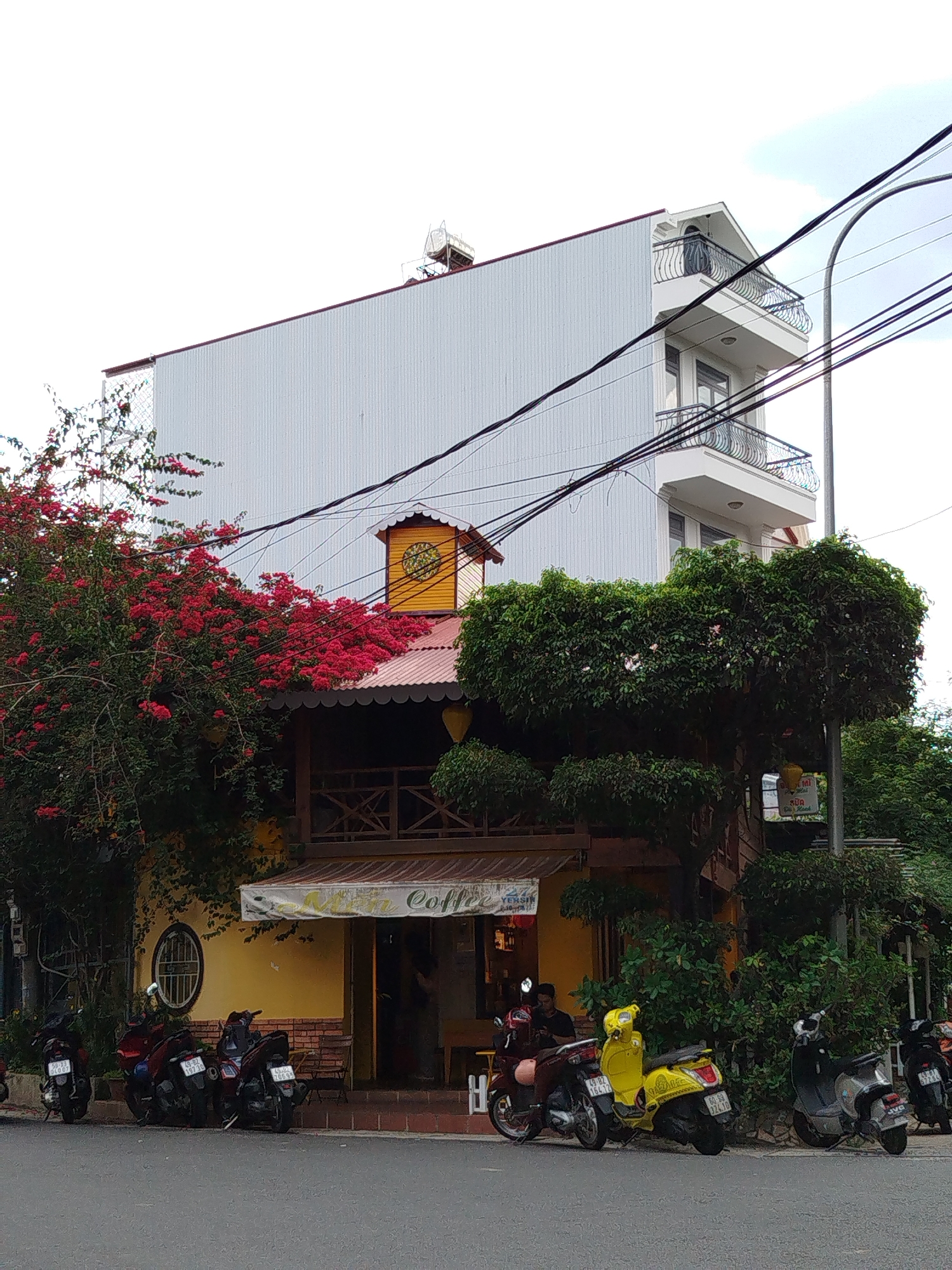 The image depicts a coffee shop in Da Lat, Vietnam, characterized by a two-story yellow and wood structure with a brown awning reading "Coffee" above the entrance. The building is adorned with lush greenery and vibrant red bougainvillea. Adjacent to the coffee shop is a taller, more modern building with white corrugated metal siding and small wrought-iron balconies. The street in front of the coffee shop is lined with parked motorcycles, including several scooters. Several people are seated near the entrance to the coffee shop. Overhead power lines cross the sky. The environment is overcast.