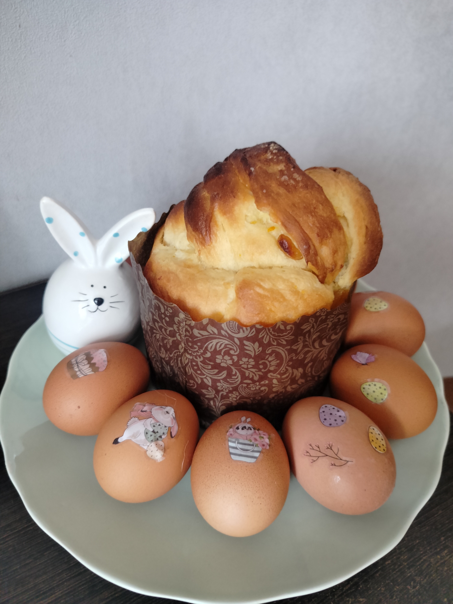 A photograph shows an Easter display featuring a braided, cylindrical bread loaf inside a brown, floral-patterned paper baking cup, surrounded by seven brown eggs decorated with Easter-themed stickers. A white ceramic bunny figurine with blue polka dots and facial features stands to the left of the bread. The arrangement is set on a light-green plate with a wavy edge, which rests on a dark-colored wooden surface. The background is a plain, light-gray wall. The location is inferred from the user query to be Chaikovsky, Russia, but there are no elements visible in the image to confirm this.