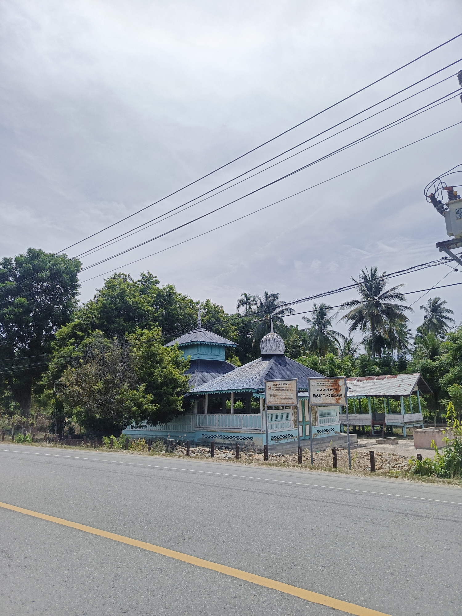 A street view shows the "Masjid Tuha Bugeng" (Bugeng Old Mosque) in Sawang, Indonesia, under a cloudy sky. The mosque features traditional blue and white architecture with a domed roof and an open veranda supported by posts. Two informational signs with Indonesian text are placed in front of the mosque, indicating "SITUS CAGAR BUDAYA" (Cultural Heritage Site). Adjacent to the mosque is an open-sided structure with a corrugated metal roof. Lush green trees, including palm trees, surround the buildings. Power lines stretch across the upper part of the frame, with an electrical transformer visible on a pole to the right. A paved road with a yellow center line runs in the foreground. The ground around the mosque is rocky and dry. There are no people visible in the image.