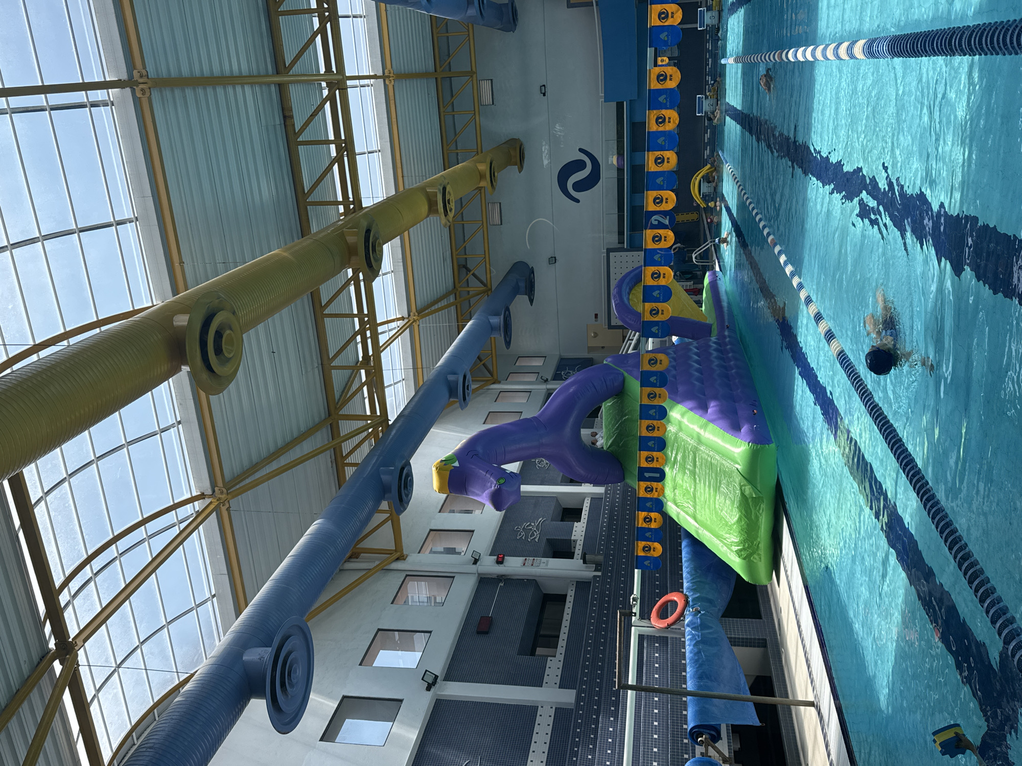 This media file captures a vibrant indoor swimming pool facility, likely located in Puebla, Mexico, from an elevated, rotated perspective. The scene is brightly lit by natural light filtering through large translucent panels in the roof, indicating it's daytime.
The main subjects are several swimmers enjoying the pool. One individual wearing a dark swim cap is visible in the foreground, partially submerged, while another swimmer is further down a lane.
The pool itself is a large, clean body of blue water, clearly marked with lane lines – some striped blue and white, others black and white. Along one side of the pool, a large, colorful inflatable obstacle course or play structure is set up, featuring distinct purple and green sections. A purple inflatable figure, resembling a dragon or monster, is part of this structure. Adjacent to the inflatables are blue and yellow banners with a repetitive logo featuring a yellow circle with an 'M' inside and partially visible text that reads "AQUA...". A red life preserver ring hangs nearby.
The setting reveals an open, modern architectural design. Overhead, prominent yellow/gold and blue cylindrical ducts run parallel across the ceiling. These ducts are suspended from a gold-colored metal framework that supports the translucent roof panels, allowing ample light into the facility. The walls are tiled in various shades of blue and white, featuring several rectangular windows. A distinctive blue swirl logo, resembling a stylized 'S' or yin-yang symbol, is prominently displayed on a back wall above the windows. The overall atmosphere suggests a recreational and active environment.