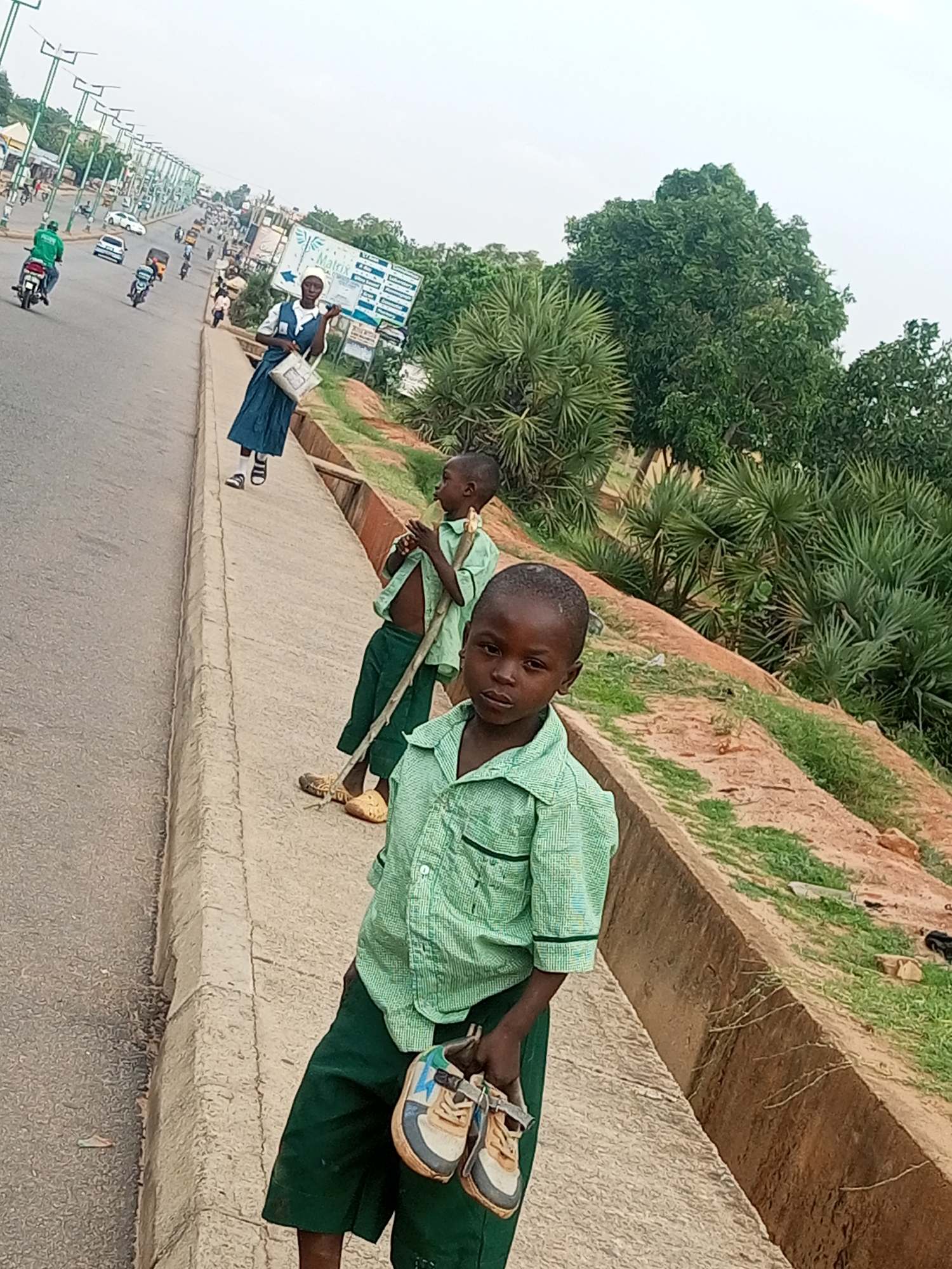 This image captures a street scene in Gombe, Nigeria. In the foreground, a young boy, wearing a green checkered shirt and dark green shorts, stands on a concrete sidewalk holding a pair of worn shoes. He is looking towards the camera. Behind him and slightly to his left, another young boy, also in a green shirt and dark green pants, is visible, holding a long stick. Further back and to the left, a young girl in a blue school uniform with a white collar and a white bag is walking on the sidewalk. The street is busy with traffic, including cars, motorcycles, and a tricycle. On the right side of the street, there is an embankment covered with sparse vegetation and some palm trees. The sky is overcast. The overall impression is of a typical day on a busy road in Gombe.