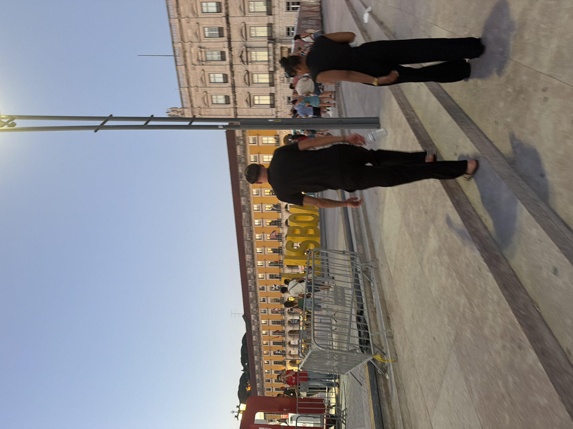 The image shows the Praça do Comércio in Lisbon, Portugal at dusk. In the foreground, a couple walks up a wide set of stone steps. The man is wearing a black t-shirt and black pants, and the woman is wearing a black sleeveless dress and black pants. They are walking away from the camera. In the mid-ground, a large yellow sign spelling out "LISBOA" is visible, with crowds of people walking behind it. Behind the sign and crowds are two large buildings. The building on the left is yellow with arched windows and balconies, while the building on the right is stone-colored with classical architectural details. A tall, modern lamppost stands to the right of the couple, casting a shadow. In the background, the sky is a clear, pale blue, transitioning to darker blue towards the top. A red structure, possibly a tram, is partially visible on the far left of the image, with a metal barrier in front of it.