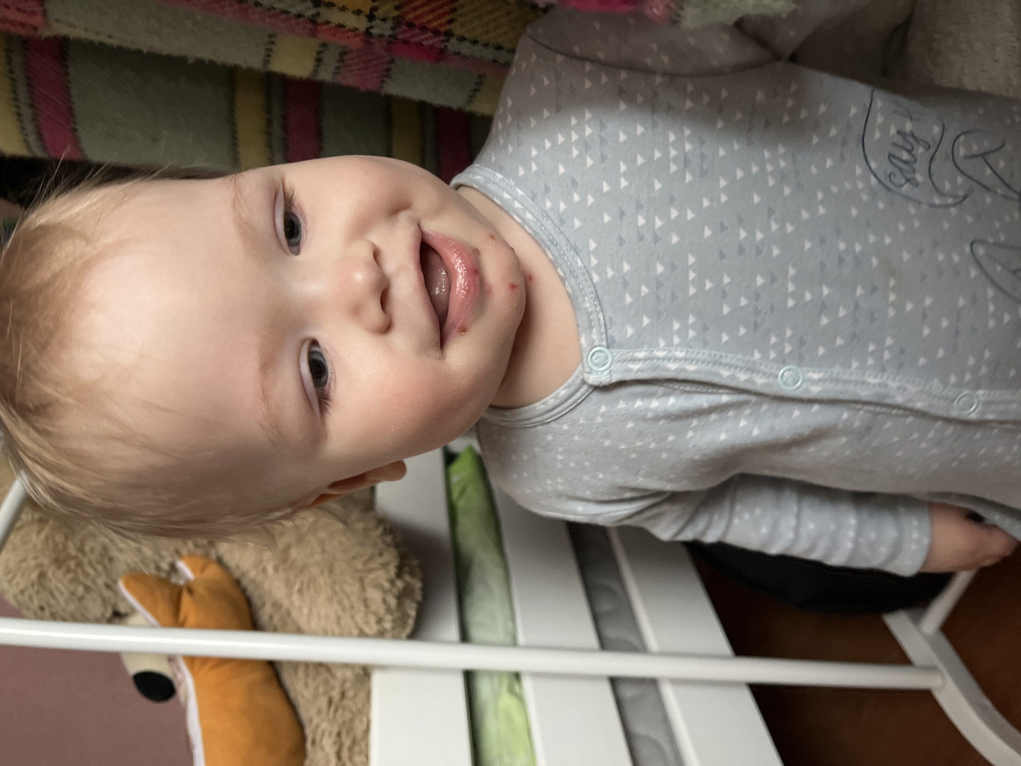 A fair-skinned baby with blonde hair is indoors, likely in a crib or playpen with white rails. The baby is wearing a gray onesie with a pattern of white triangles and a cartoon image on the chest. The baby has a slightly open mouth, with visible red marks around the lips and mouth area. The background includes a multicolored blanket or fabric and a brown teddy bear with a fox-like plush toy partially visible. The location of the image is not recognizable as Chaikovsky, Russia, or identifiable as any specific location from the visual information provided.