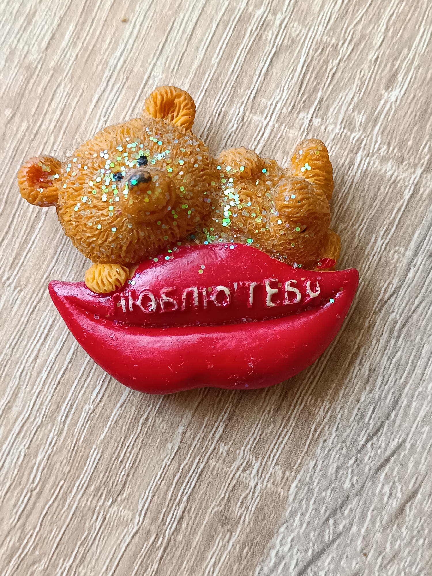 A close-up, overhead shot displays a small, glitter-covered teddy bear figurine lying on its back atop a pair of bright red, glossy lips. The bear is light brown and adorned with iridescent glitter that catches the light. Its eyes are black, and its paws are resting on the lips. The lips appear to be a separate embellishment, with the Russian text "ЛЮБЛЮ ТЕБЯ" (I love you) embossed in white lettering across the bottom lip. The entire item is placed on a light brown, wood-grained surface, possibly a table or desk. The location is not specified in the image.