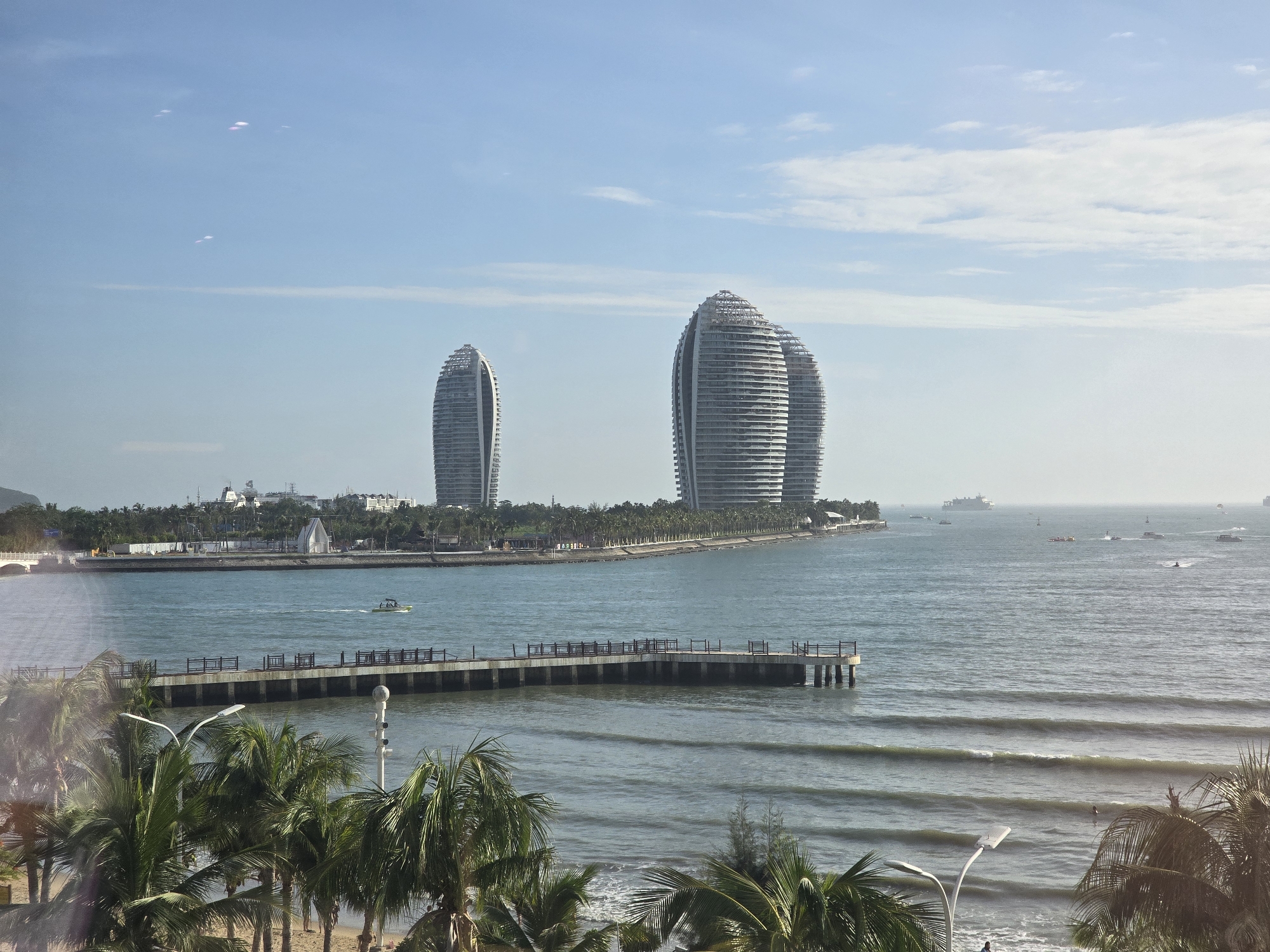 The image captures a waterfront scene in the Tianya District, China, dominated by the distinctive "Coconut-shaped" hotels that resemble large, segmented shells. These modern architectural marvels are situated on a peninsula jutting into the ocean. In the foreground, a sandy beach is lined with lush palm trees, their fronds partially obscuring the view. A pier extends into the calm, blue ocean, with several small boats dotting the water. Further out, larger vessels, including what appears to be a cargo ship and several smaller boats, are visible on the horizon under a clear, pale blue sky with sparse clouds. The water exhibits gentle waves, and a small boat is actively moving on the sea near the pier. The overall impression is one of a modern coastal resort area.