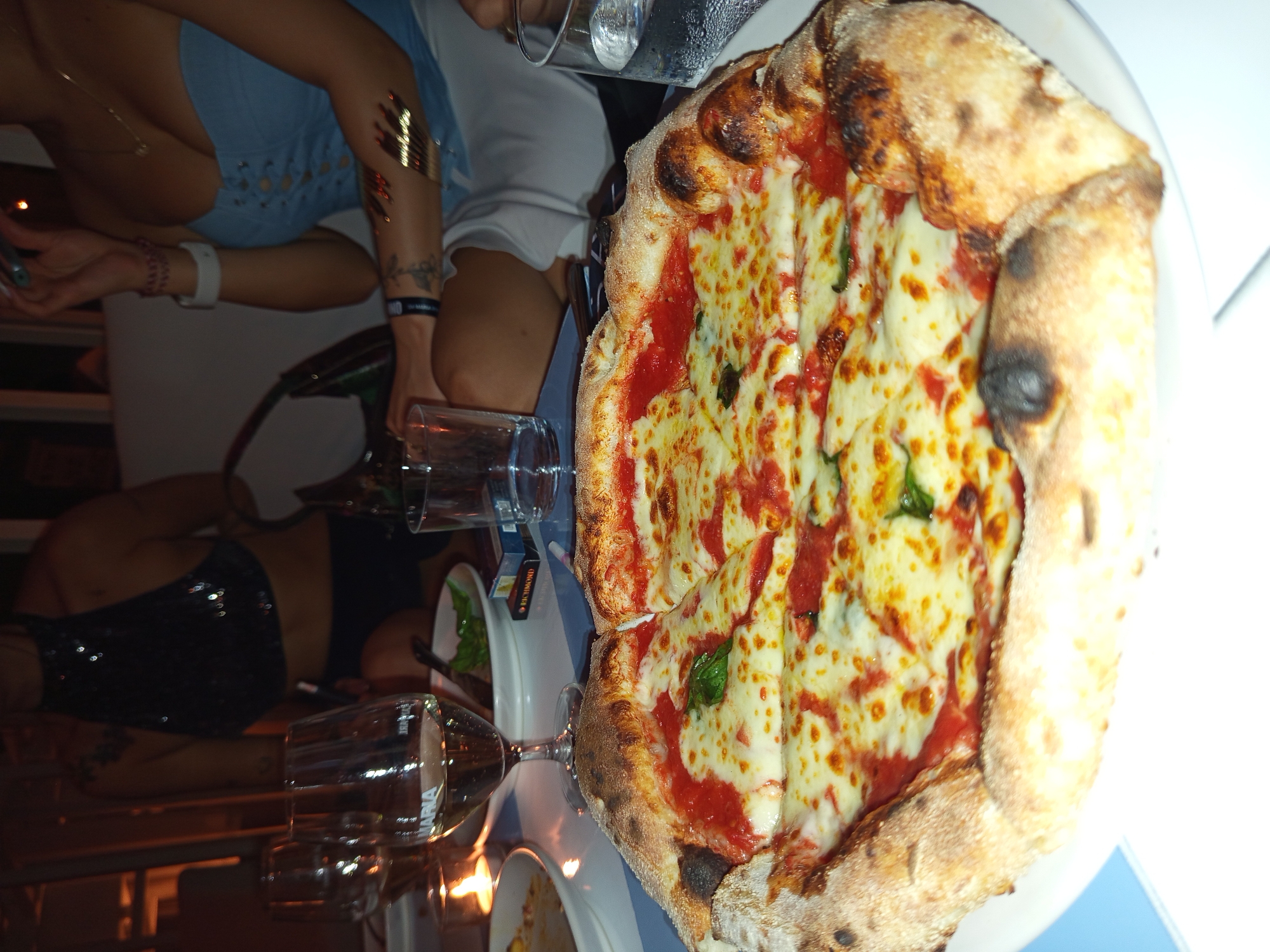 The image shows a group of people at a table, possibly dining. A pizza with a thick crust, topped with tomato sauce, cheese, and basil, is the primary focus, placed on a blue-striped plate. There are also glasses and other plates with food around it. The image was taken in North Kuta, Indonesia.