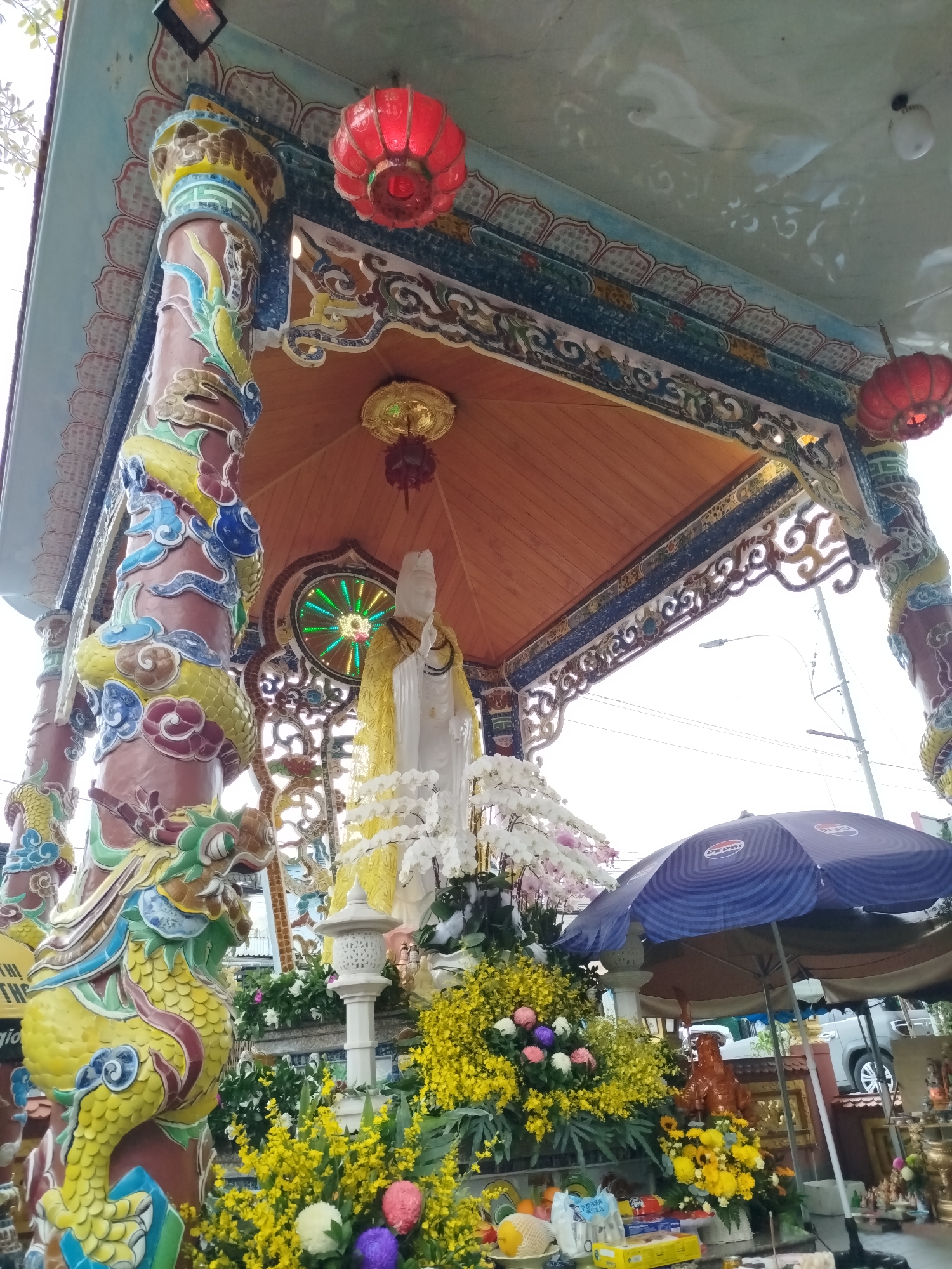 This image captures a vibrant and ornate outdoor shrine, likely in Da Lat, Vietnam. The central focus is a tall, white statue, possibly of Quan Yin (Guan Yin), draped in a yellow garment, standing beneath an elaborate canopy. The statue is surrounded by numerous floral offerings, including abundant yellow orchids, elegant white orchids, and various other colorful flowers, suggesting a devotional or celebratory context.
The canopy is supported by intricately carved pillars adorned with colorful dragon motifs in shades of yellow, blue, green, red, and brown. The canopy itself features a wooden underside and decorative, carved edges. Red paper lanterns hang from the canopy, adding to the cultural aesthetic.
On the right side of the image, a large blue umbrella bearing the "PEPSI" logo shades an area containing further offerings, such as fresh fruits (one netted), packaged goods, and smaller statues or religious items. A white, ornate stone lantern stands near the base of the main statue. In the background, elements of an urban setting are visible, including power lines, a street light, and a modern car. A partial yellow sign on the far left displays the text "THI THÉ gio". The lighting suggests it is daytime, providing a clear view of the shrine's rich details and the surrounding devotional setup.