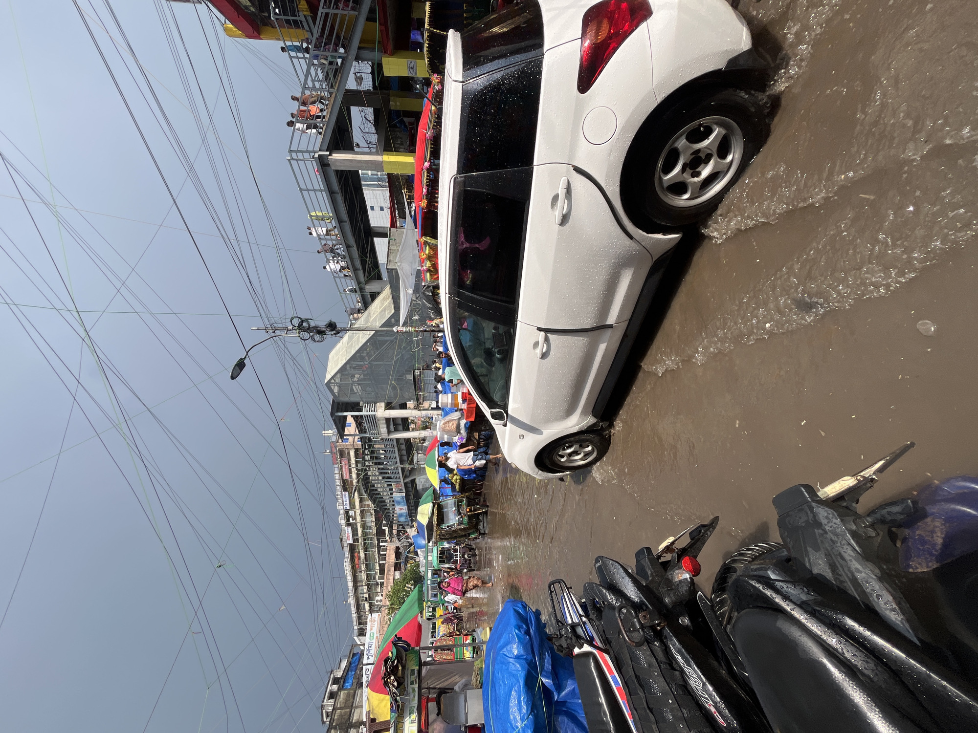 The scene is a flooded street in Dhaka, Bangladesh. A white hatchback car is partially submerged in the murky water. Parked motorcycles are also partially underwater in the foreground. People are wading through the floodwaters in the background. The scene features buildings and overhead power lines. Vendors under colorful umbrellas attempt to sell goods. A metal pedestrian walkway is visible. The sky is overcast and grey.
