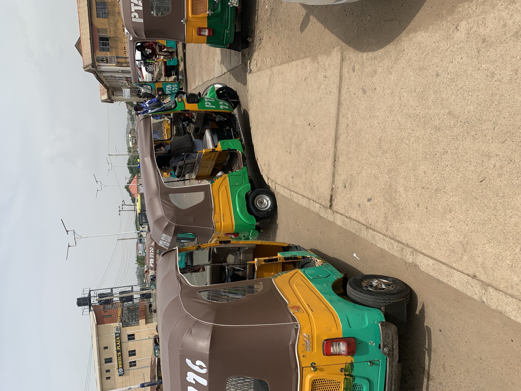 The image shows a street scene in New Stadium, Nigeria, with a row of auto-rickshaws (tuk-tuks) parked along the side of the road. The tuk-tuks are painted in a combination of green, yellow, and brown. They display "PTA" followed by numbers, which may be registration or identification codes. The vehicles are parked close together, indicating a designated area or taxi rank. In the background are buildings, including one named "Wheel's Plaza" with advertisements for "GEG WHEELS." One of the tuk-tuks has text "DO NOT URINATE P" visible on its body. There are people visible inside one of the tuk-tuks. The day appears to be bright.