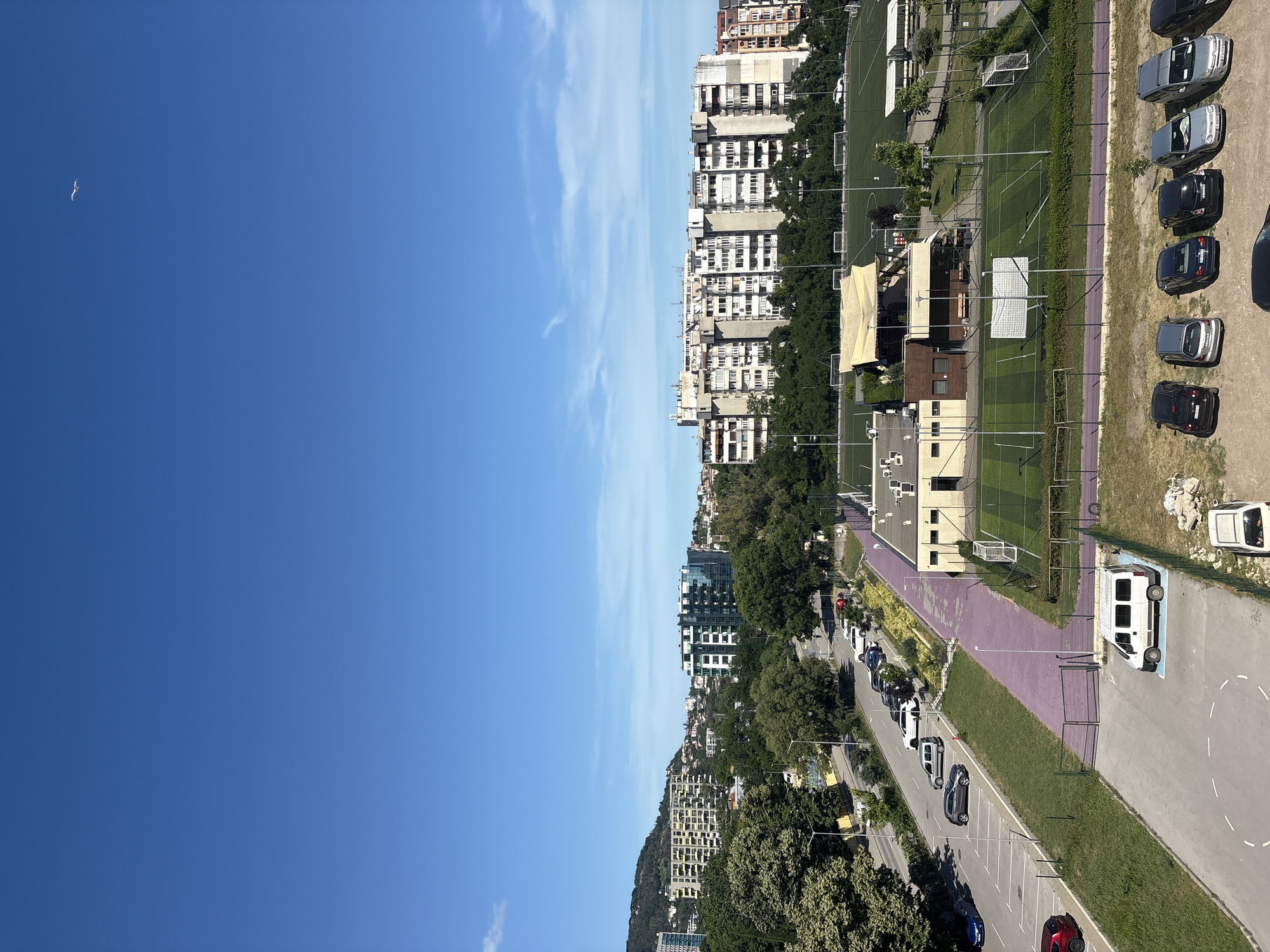 This is a high-angle, full-shot, color photograph taken on a clear, sunny day. The image is oriented vertically, with the sky occupying the left half of the frame and a cityscape and sports facilities on the right. The sky is a vibrant, cloudless blue, with a single white bird visible in the upper left quadrant.
The right side of the image displays a dense urban environment with numerous apartment buildings. These buildings appear to be of varying heights and architectural styles, with many featuring balconies and numerous windows. Adjacent to the buildings is a sports complex featuring at least two green soccer fields with white goalposts. A purple running track runs along the edge of one of the fields.
A parking lot is visible in the foreground, with several cars parked in designated spots. These include a white van, several dark-colored cars, and a couple of silver cars. The ground in the parking lot is a mix of asphalt and gravel. Trees and other greenery are interspersed throughout the scene, particularly between the buildings and the sports fields, and along the roadside.
The lighting suggests it is daytime, likely late morning or early afternoon, given the bright sunlight and distinct shadows. There are no people visible in the image. The overall impression is of a developed urban area with recreational facilities, set against a backdrop of blue sky.