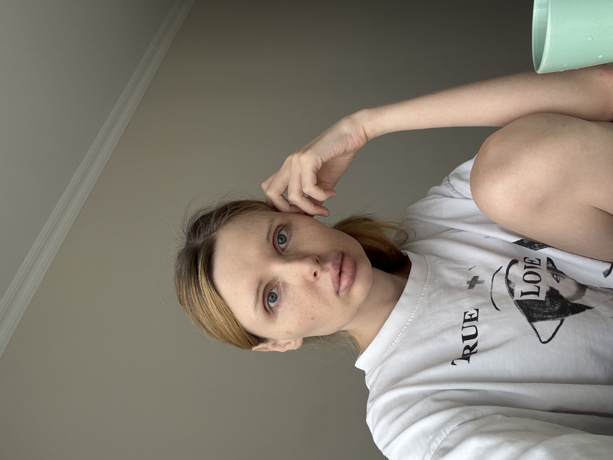 A Caucasian woman with blonde hair pulled back into a ponytail, fair skin with freckles, and light blue eyes is shown from the chest up, looking directly at the camera. She is wearing a white t-shirt with black lettering that appears to read "TRUE LOVE." Her right hand is resting on her forehead, and her left arm is bent with her hand resting on her knee. She is positioned indoors with a plain, light-colored wall and ceiling visible in the background. To the right of the frame, a light teal mug with water droplets on the outside is partially visible. The lighting suggests a natural light source from the left. The overall scene appears to be casual and unposed, possibly taken in a home environment. There are no visible indications of the location within a city.