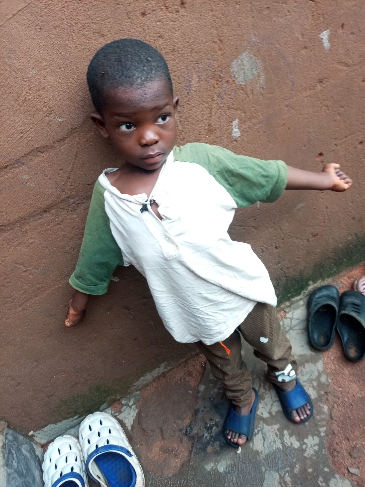 A young Black boy is leaning against a textured brown wall in an outdoor setting. He is wearing a white t-shirt with green sleeves, brown pants, and blue sandals. His right arm is extended outwards, and his left arm is bent, with his hand resting on his hip. In the foreground, to the left, a pair of white sandals with blue accents are visible. To the right, a pair of black sandals are placed on the ground. The ground is a mixture of dirt and concrete. The overall scene suggests a casual, everyday moment.