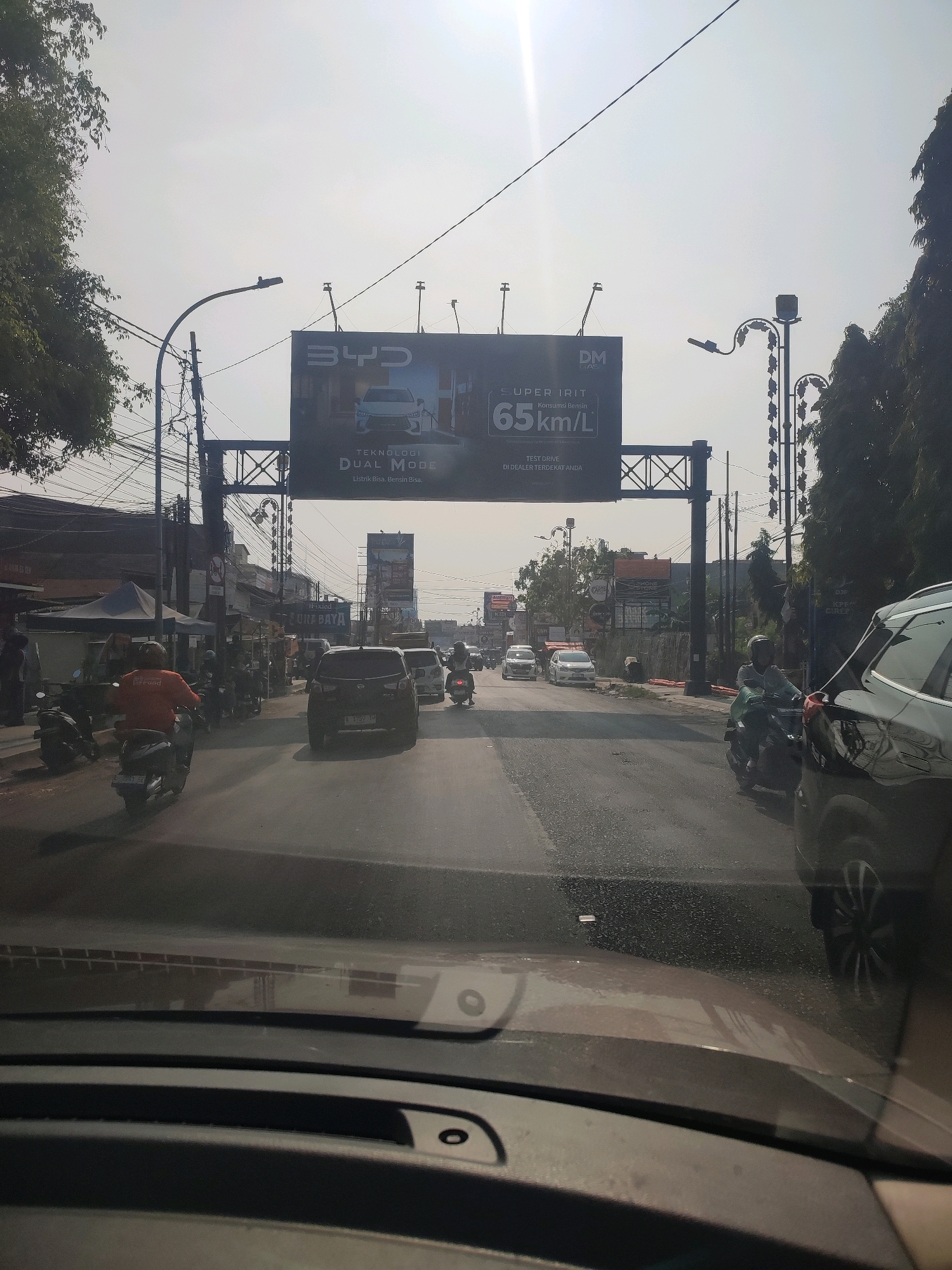 The image depicts a street scene in Sukapura, Indonesia, under a bright, overcast sky. A large billboard advertising BYD Dual Mode technology with a fuel efficiency of 65 km/L is prominently displayed above the road. The billboard also encourages a "TEST DRIVE DI DEALER TERDEKAT ANDA."
The road is filled with various vehicles, including cars and motorcycles. In the foreground, the perspective suggests the photo is taken from inside a car, with the dashboard visible. Several motorcycles are seen on the left side of the road, with one rider wearing an orange shirt. A black SUV is in the right lane, partially obscuring a white car. Further down the road, more vehicles, including cars and motorcycles, are navigating the traffic.
The surrounding environment includes buildings, trees, and utility poles with a complex network of wires. A few tents are visible on the left side of the road, suggesting commercial activity. The overall atmosphere is that of a busy urban street during daylight hours.