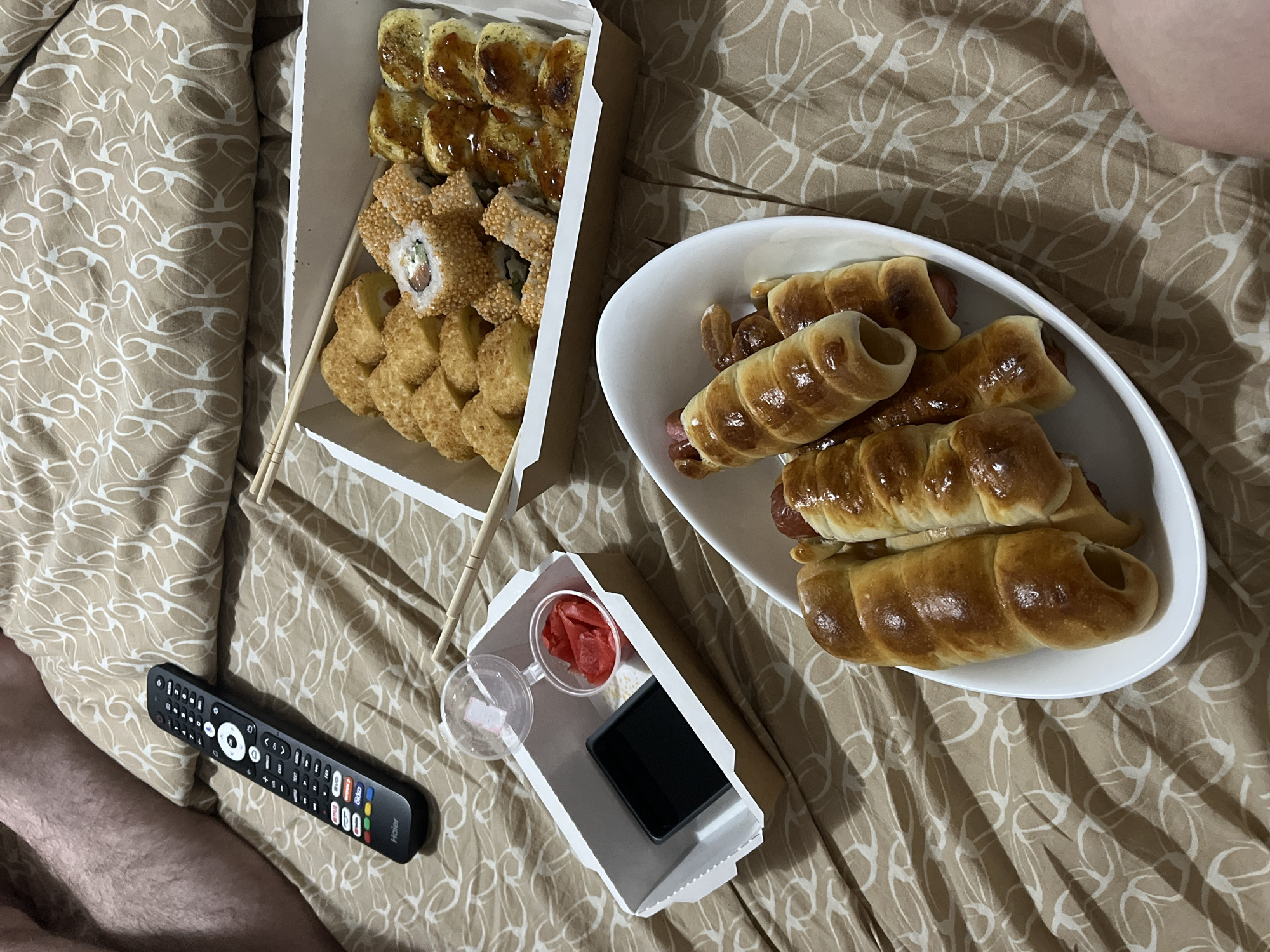 The image shows a meal laid out on a bed. There is a white cardboard container filled with sushi rolls, including tempura, sesame-coated, and eel varieties. Beside the sushi is a white oval-shaped dish containing several "pigs in a blanket", sausages wrapped in baked dough. A smaller white cardboard box holds condiments: a plastic container of pickled ginger and what appears to be a cell phone. Chopsticks are present alongside the sushi. A black remote control is lying on the bed beside the smaller white box. The bed has a patterned beige and white duvet cover. The image may be taken in Chaikovsky, Russia.