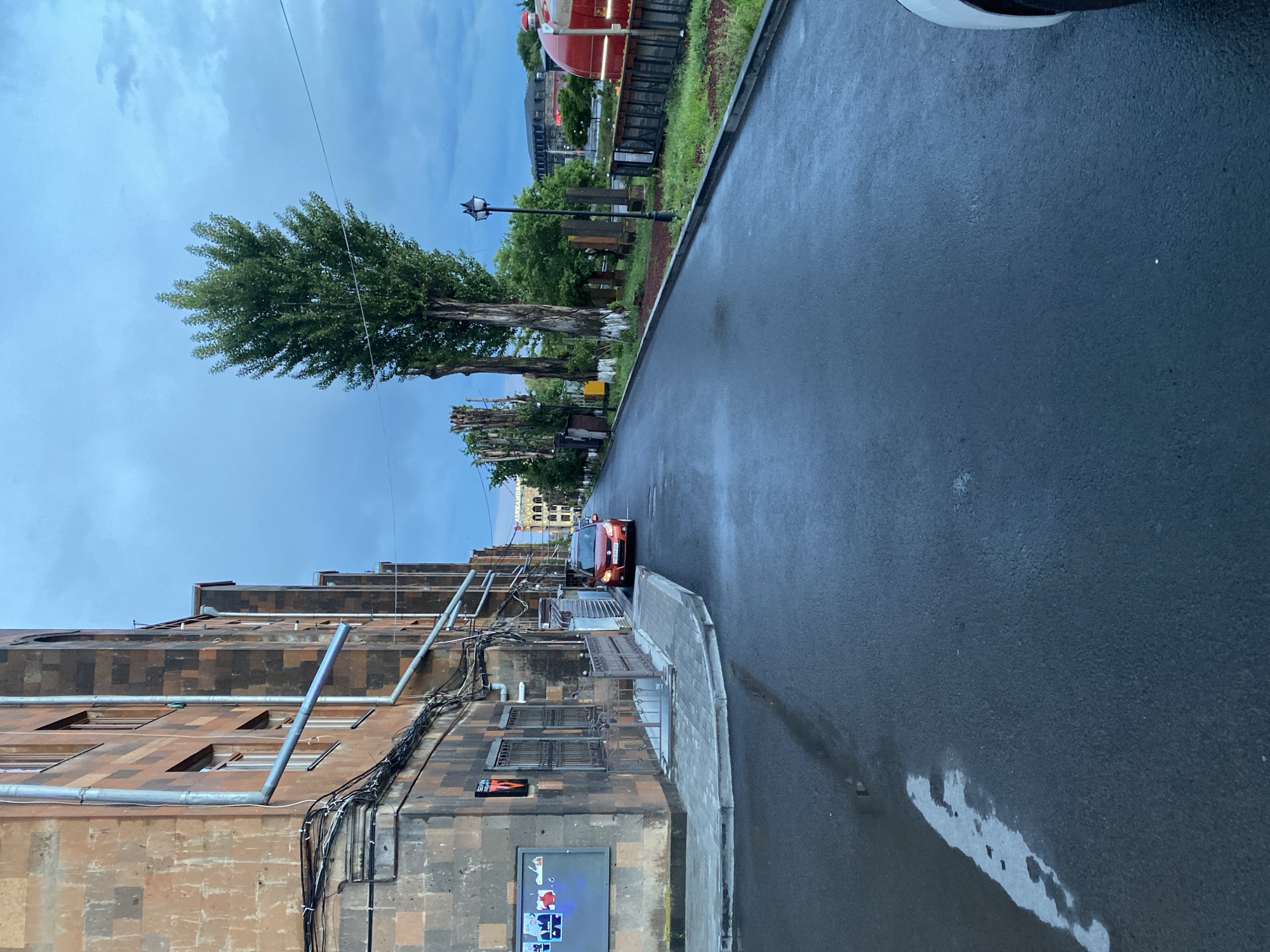 The image depicts an exterior view of a street in Gyumri, Armenia, likely in the evening or during overcast conditions, as indicated by the muted light and dark blue sky. A multi-story building with a reddish-brown stone facade runs along the left side of the frame, characterized by numerous external pipes and wiring. Adjacent to the building is a sidewalk with a low metal railing. A red sedan is parked on the wet asphalt road. The road stretches into the distance, flanked by trees and a grassy area on the right. In the background, several structures are visible, including a building with arched windows and a red, train-like structure with lights. The presence of puddles on the road suggests recent rainfall. The overall atmosphere is urban and somewhat industrial.