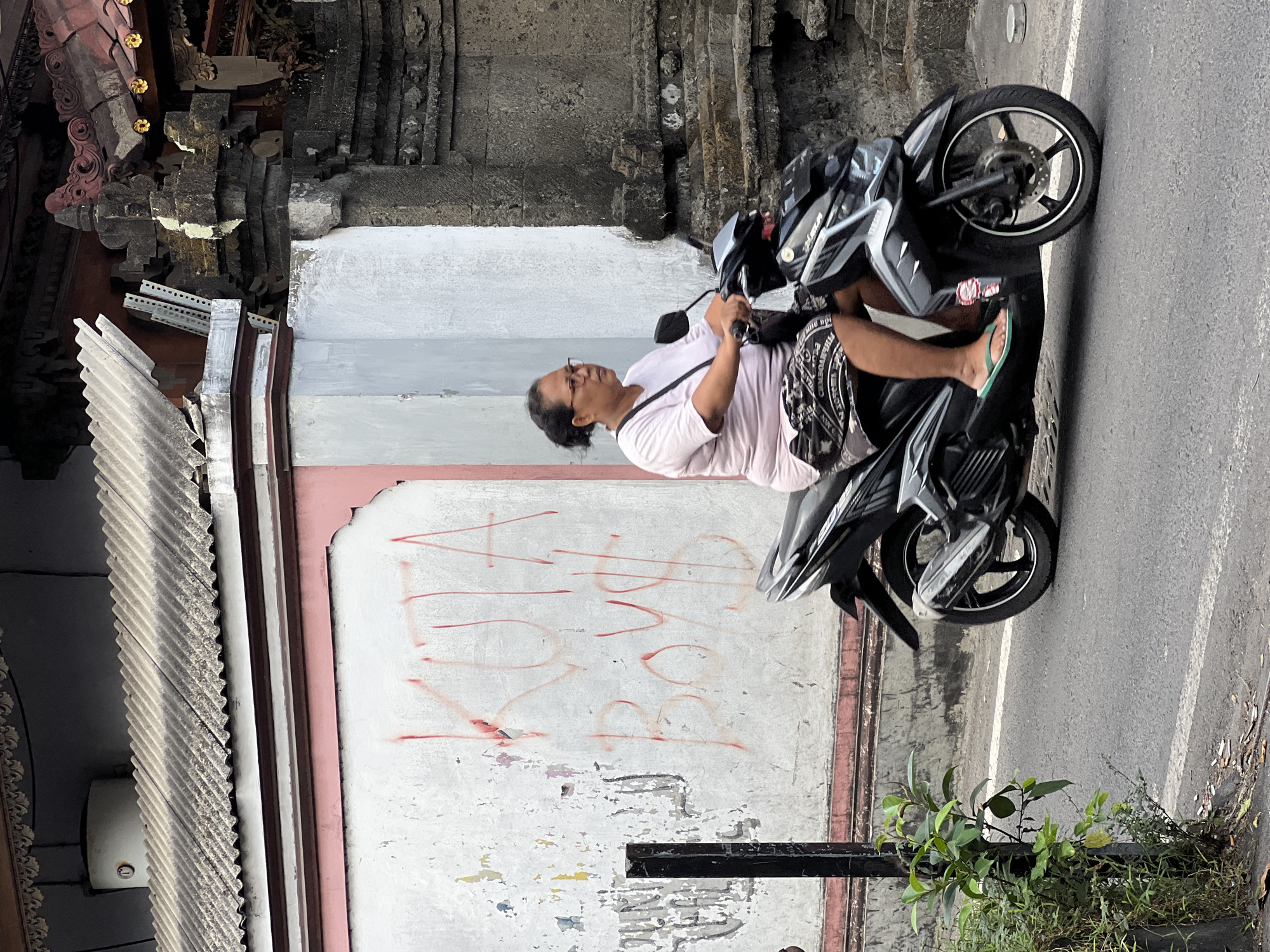A woman rides a black scooter on a street in Kuta, Indonesia. She is wearing a light pink t-shirt, a patterned skirt, and green flip-flops. The wall behind her is white with pink trim and red graffiti that reads "KUTA BOYS". To the right of the wall is a stone structure, possibly part of a temple. Green foliage is visible in the foreground on the left side of the image.