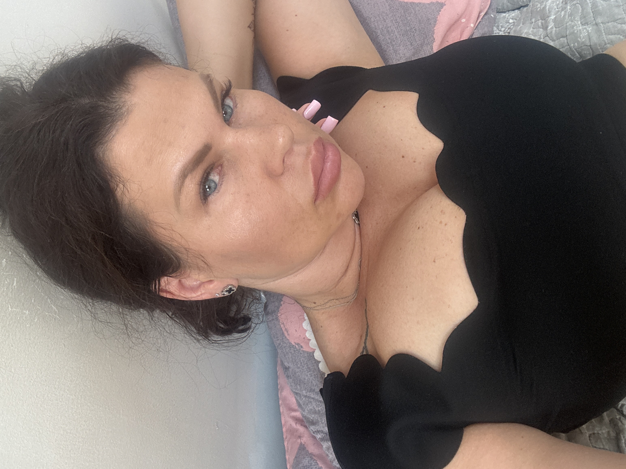 The image is a close-up, overhead selfie of a woman lying down. She has dark hair, blue eyes, and light skin with freckles. She is wearing a black top with a scalloped neckline, a silver necklace, and an earring in her left ear. Her right arm is behind her head, and her left arm is resting on her chest. The background appears to be a bed with a patterned comforter and a white wall. There is no indication of the city of Katowice, Poland, or any specific urban environment. The image is a personal photograph, likely taken in a private residence.