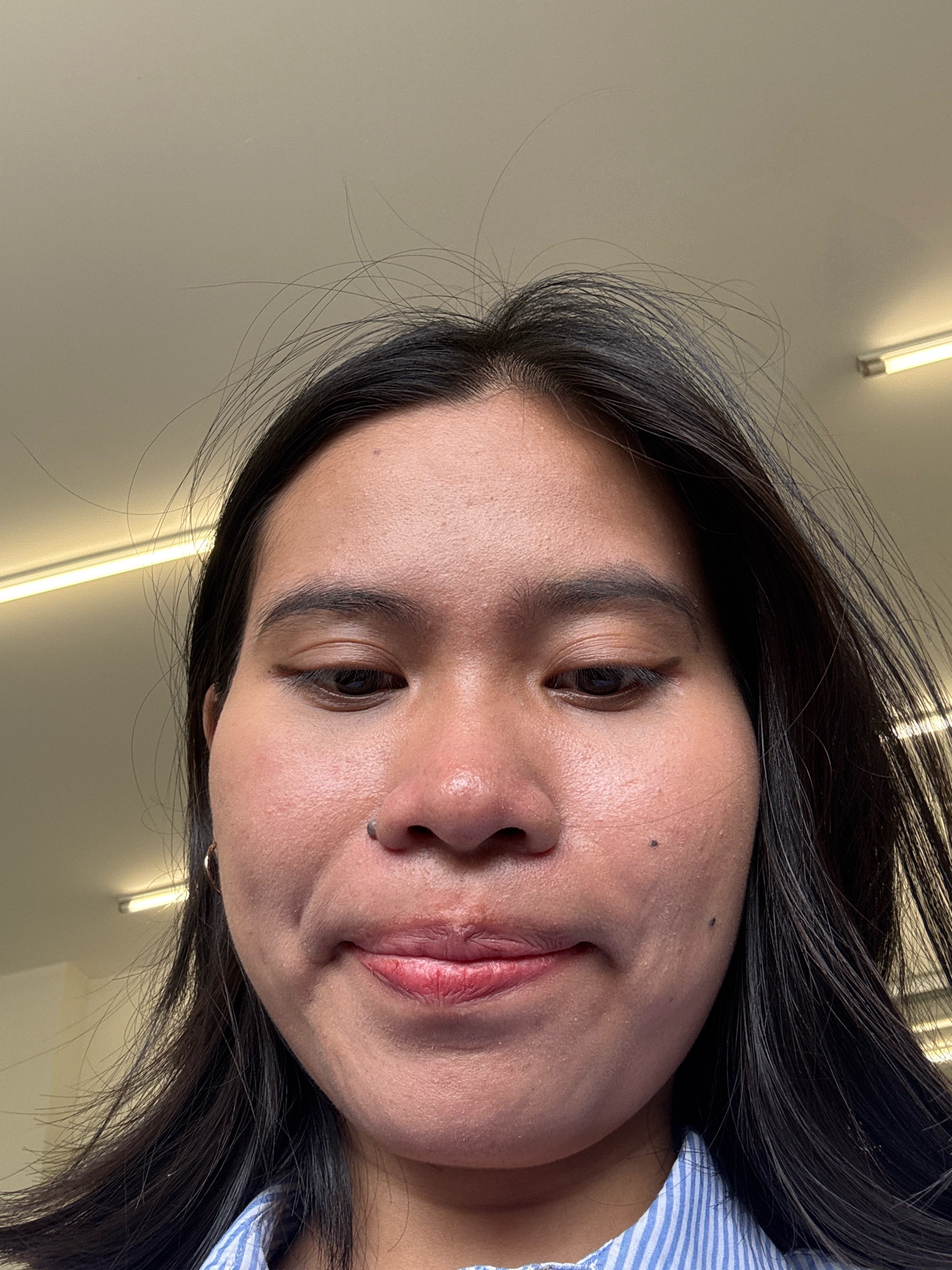 A close-up, low-angle shot shows a young woman's face from just above her eyebrows down to her chin. Her dark hair is spread out around her face. She has a light complexion with some visible pores and blemishes. Her eyes are looking downwards. She is wearing a blue and white striped shirt. The background appears to be an indoor setting with ceiling lights visible. The image is taken in East Jakarta, Indonesia.