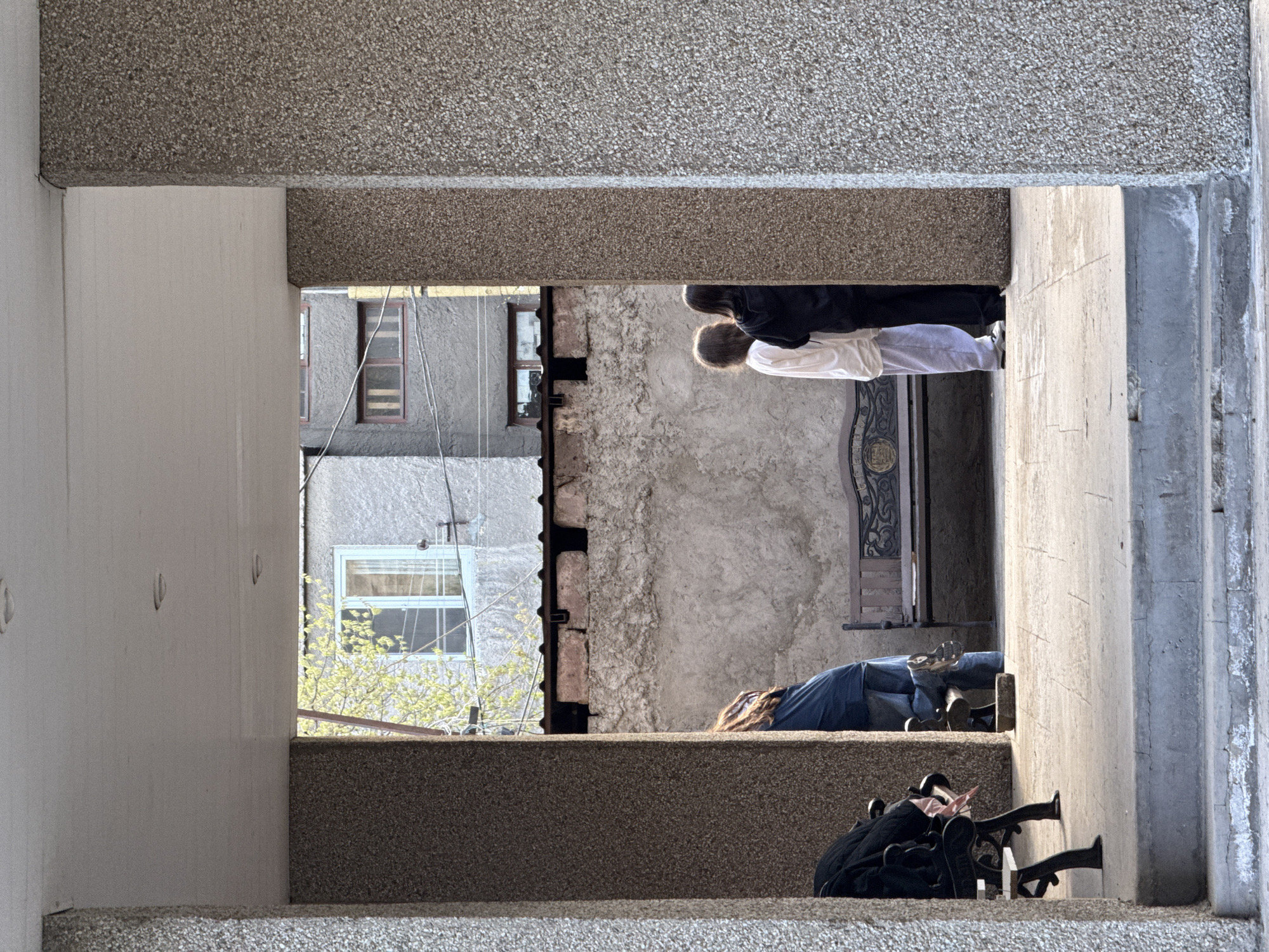 A low-angle, medium shot is taken through a concrete architectural element, capturing an outdoor scene in Yerevan, Armenia. The photograph is oriented sideways, with what would be the ground on the bottom and the sky at the top in a standard view.
People are visible in the scene. Two people are on a ledge, their torsos and heads visible; one wears white and the other wears black. Below and to the left, another individual lies on a concrete ledge, dressed in a blue top and pants. A black bag and bicycle wheels are situated on the concrete surface beneath them.
In the background, a gray building with windows is partially visible. A concrete wall with attached metal braces is on the center of the view.