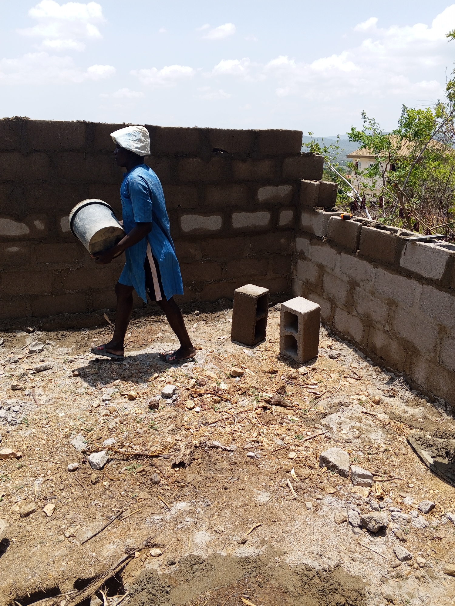 An exterior shot in Lokoja, Nigeria, depicts a construction site. A man is carrying a bucket in the foreground, wearing a blue robe over shorts, sandals, and a white head covering. Behind him is a partially constructed block wall, with two hollow concrete blocks resting on the dirt ground near the wall's corner. The ground is covered with dirt, small rocks, and bits of debris. In the background is a distant landscape with greenery and a building partially visible to the right of the wall. The sky is partly cloudy.