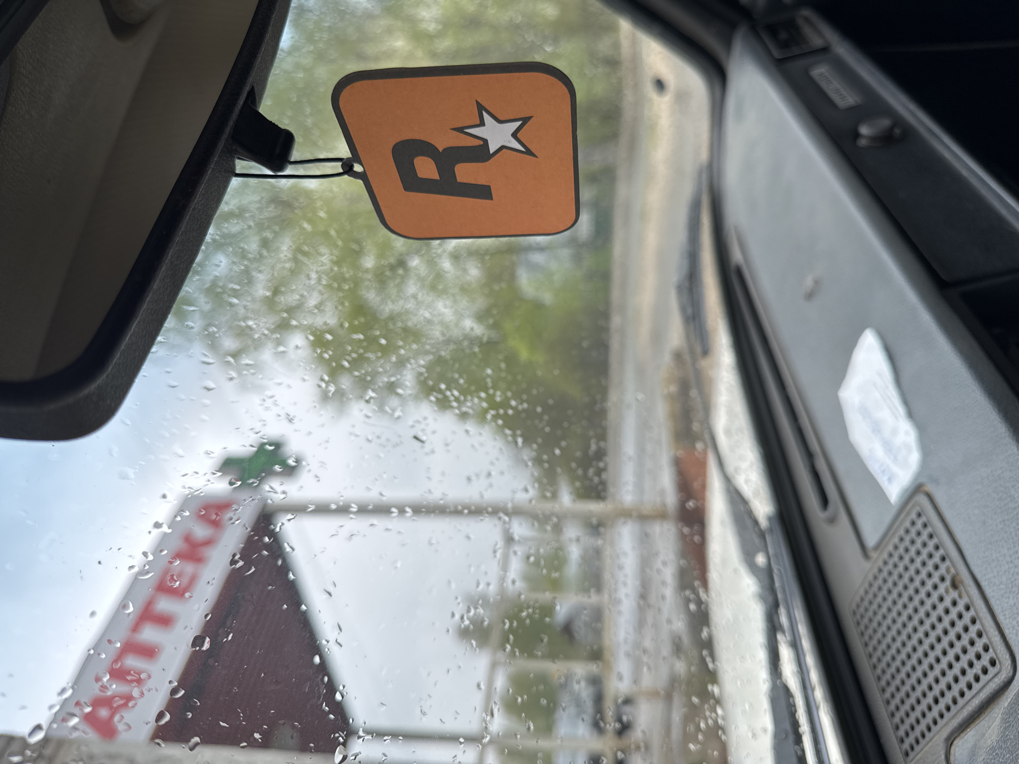 The image shows a view from inside a vehicle, likely a car, on a rainy day. Raindrops are visible on the windshield. An air freshener with the Rockstar Games logo hangs from the rearview mirror. Through the windshield, a building with a sign that reads "Apteka" (pharmacy) is visible. The scene is likely in Chuprino, Russia, given the signage in Russian. The focus is on the interior of the vehicle and the view of the pharmacy outside.