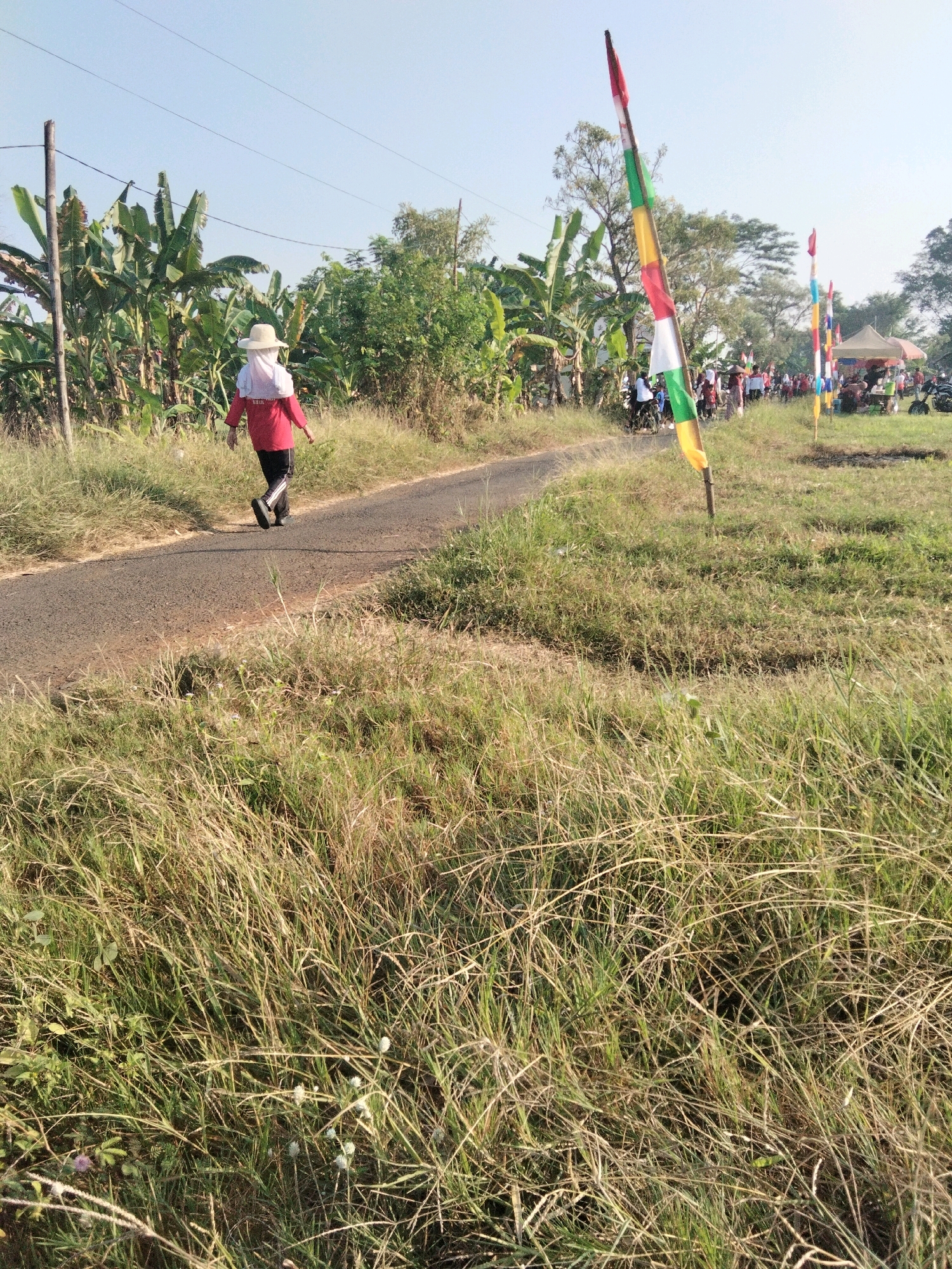 A person walks on a paved road lined with dry grass and banana trees in Ponowareng, Indonesia. The individual is wearing a red long-sleeved shirt, black pants, a white headscarf, and a straw hat. In the background, several people are gathered near tents and stalls, with colorful flags on poles decorating the area. The sky is clear and blue, indicating it is daytime.