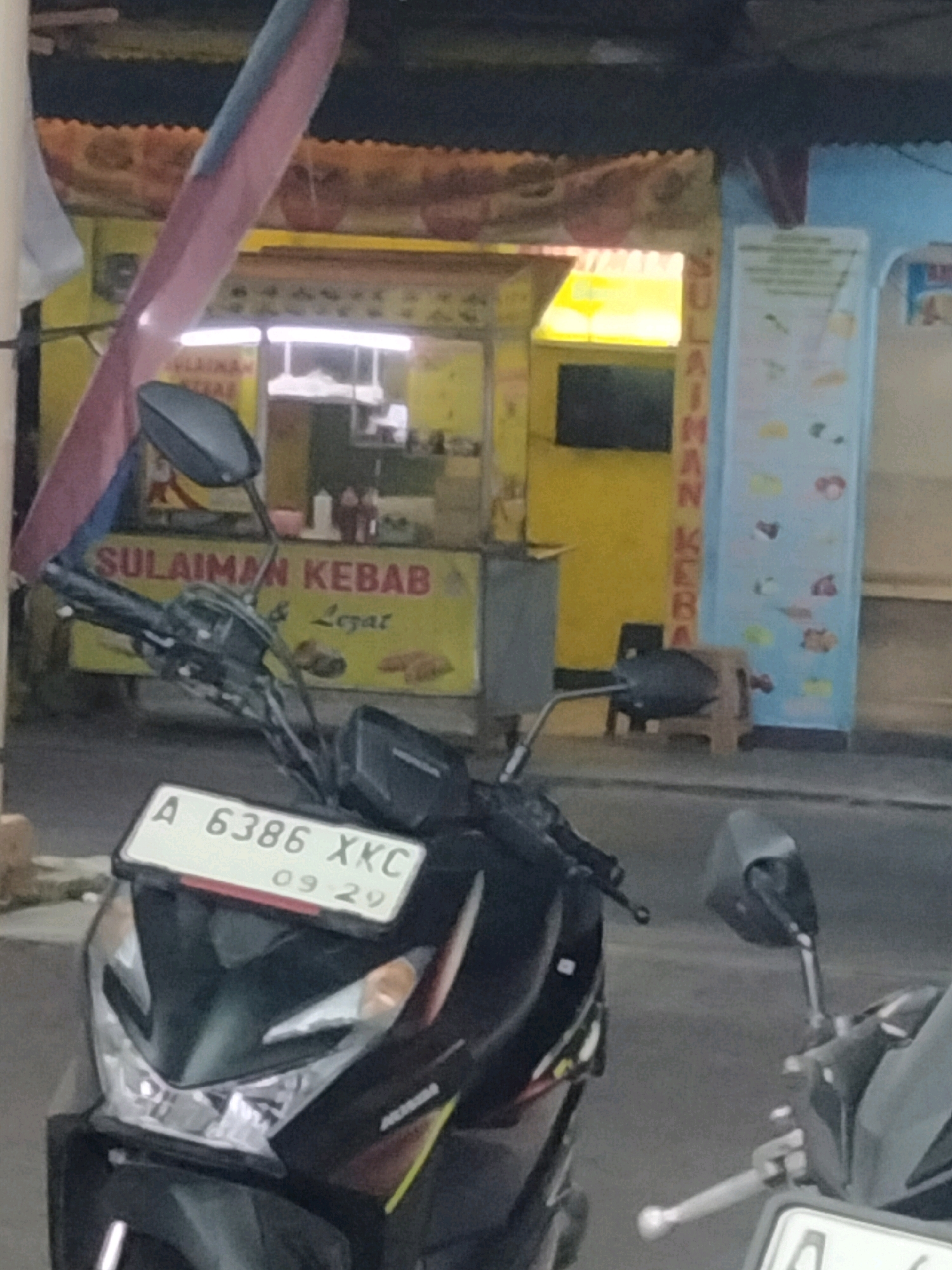 The image shows a street scene in Pisangan Jaya, Indonesia, likely a commercial area given the presence of food stalls and signage. In the foreground, the front of a black motorcycle is visible, featuring a white license plate with the characters "A 6386 XKC" and the date "09 29". The motorcycle's headlight and front fairing are clear. Behind the motorcycle, a "SULAIMAN KEBAB & Lezat" food stall is evident, with a yellow facade and red lettering. A vertical sign with a list of items, possibly food options, is displayed on a light blue background to the right of the kebab stall. The background suggests an outdoor setting with some roofing structures and what appears to be a street or pavement. The overall lighting suggests it is daytime, though potentially overcast or in shade. There are no people clearly visible in the image.