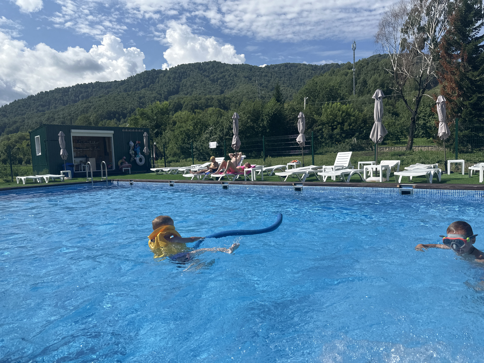 A sunny day at an outdoor swimming pool facility in Danilovka, Altai Krai, Russia, features children swimming and adults relaxing. In the foreground, two boys are in the bright blue water. The boy on the left, wearing a yellow life jacket, is swimming while holding a blue pool noodle. The boy on the right, wearing a snorkeling mask, is also swimming. In the background, several white lounge chairs are arranged on a grassy area bordering the pool, with some occupied by sunbathers. A green building, possibly a changing room or snack bar, is situated behind the lounge chairs. The facility is nestled against a backdrop of a lush, tree-covered mountainside under a partly cloudy sky.