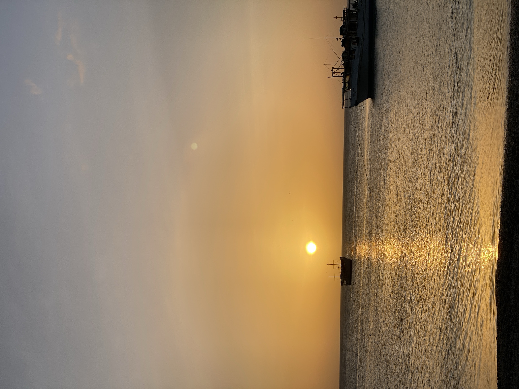 The image shows a sunset over the sea in Juan Griego, Venezuela. The sun is low in the sky, creating a golden glow. A large ship is visible in the top right of the image, and a smaller boat is visible near the sun. The water reflects the sunlight, creating a shimmering effect. The sky is a blend of gray, orange, and yellow hues. A dark shoreline is located on the right.