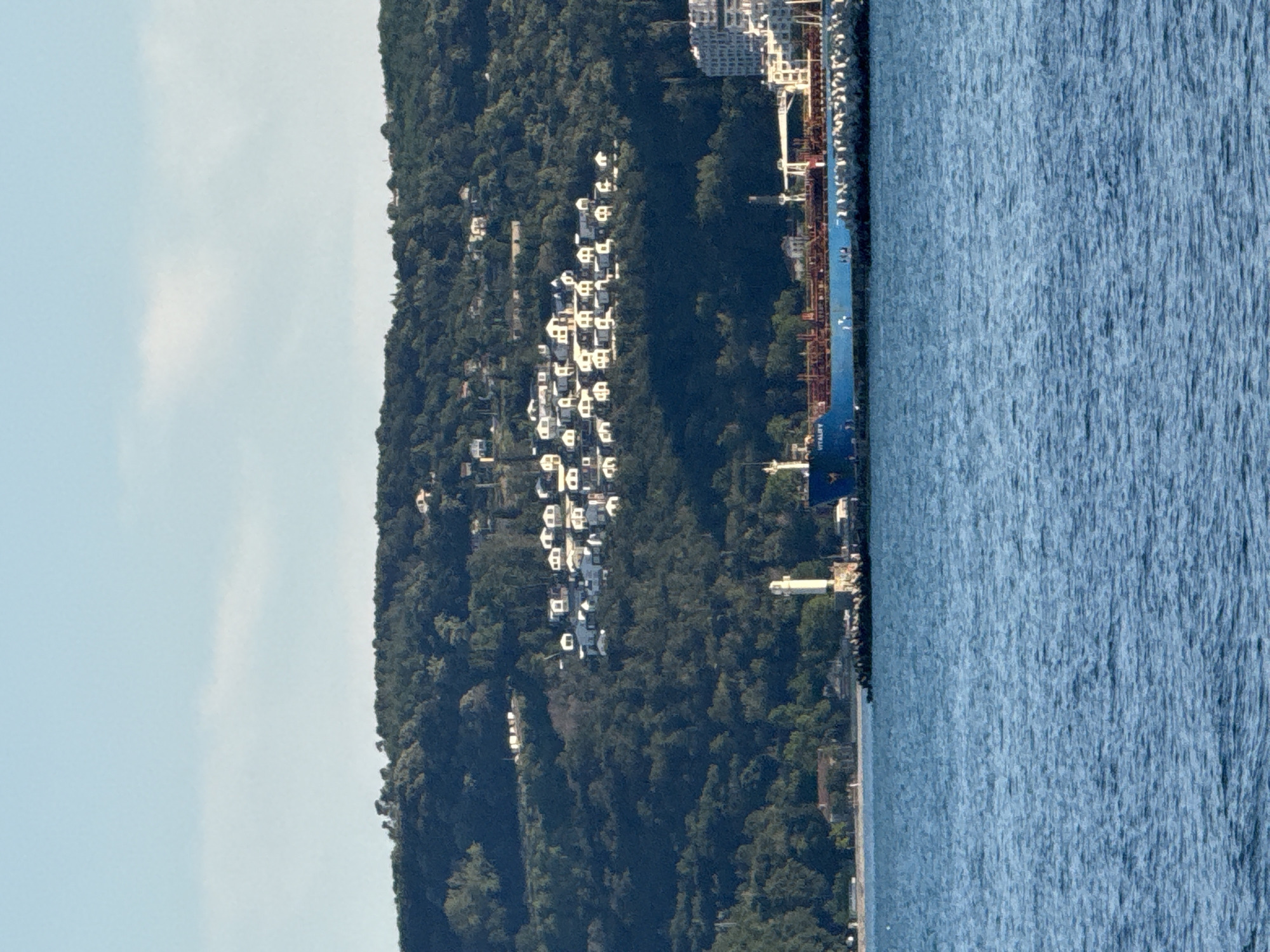 The image shows a view of a bay with a wooded hill covered in white houses in the background. A large blue tanker ship is docked in the foreground, with a breakwater extending into the water. A lighthouse is visible on the breakwater. The sky is clear and blue with a few wispy clouds. The water is calm and blue. The overall scene depicts a coastal town with a working harbor.