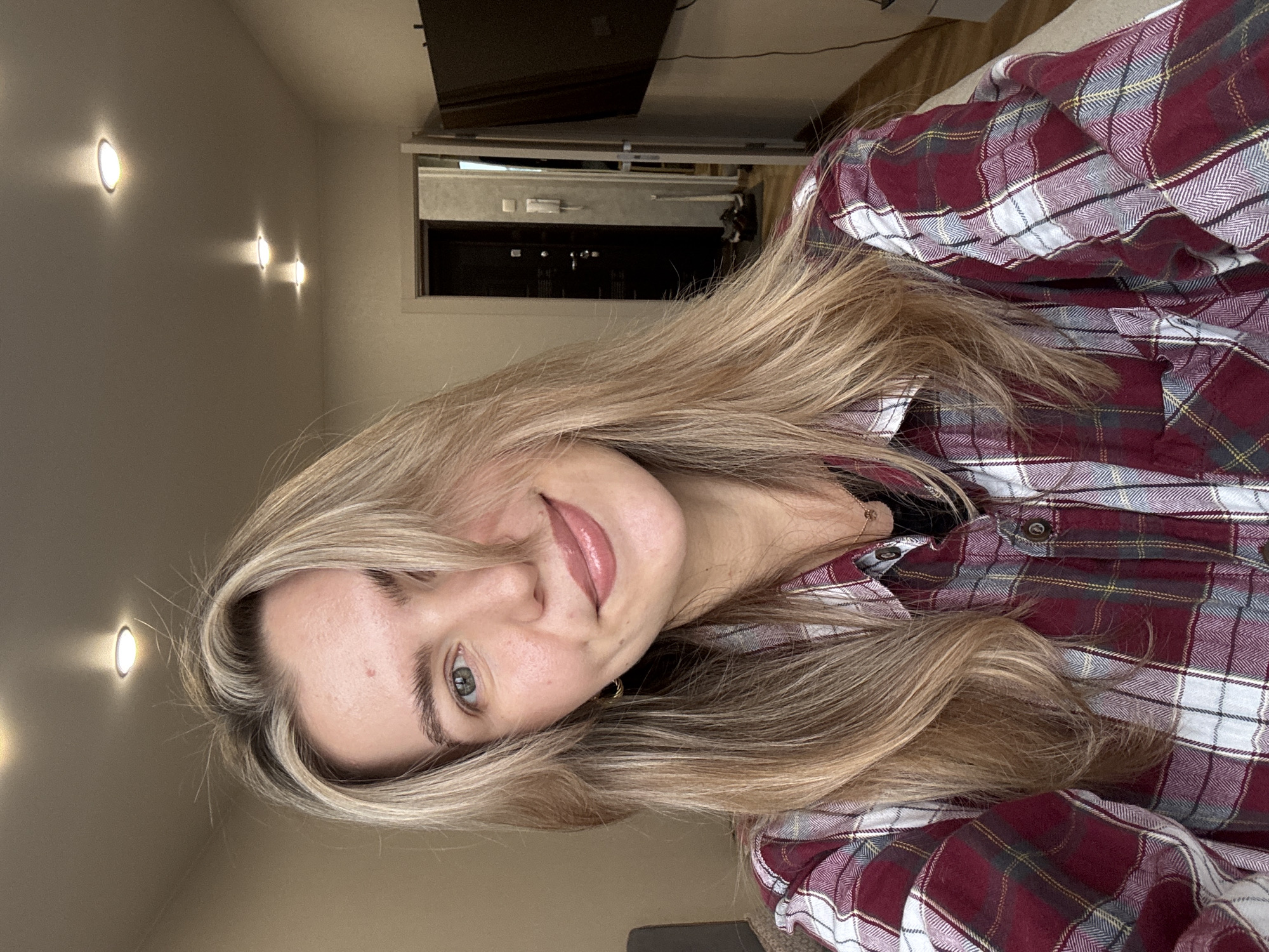 A casual, close-up portrait shows a young woman looking directly at the camera with a gentle, slight smile. She has long, blonde hair, some strands of which fall across her face. Her eyes appear to be a light color, possibly blue or green, and she has subtle makeup, including glossy lips. She is wearing a red, white, and dark blue plaid button-down shirt.
The setting is indoors. Behind her to the left, a light-colored wall features three circular recessed wall lights, emitting a warm glow. To her right, a dark, rectangular screen (possibly a television) is mounted on the wall, and below it, a dark opening with some visible metallic components suggests shelving or a media console. The lighting indicates an indoor setting, likely evening or a cozily lit space. No text is visible in the image.