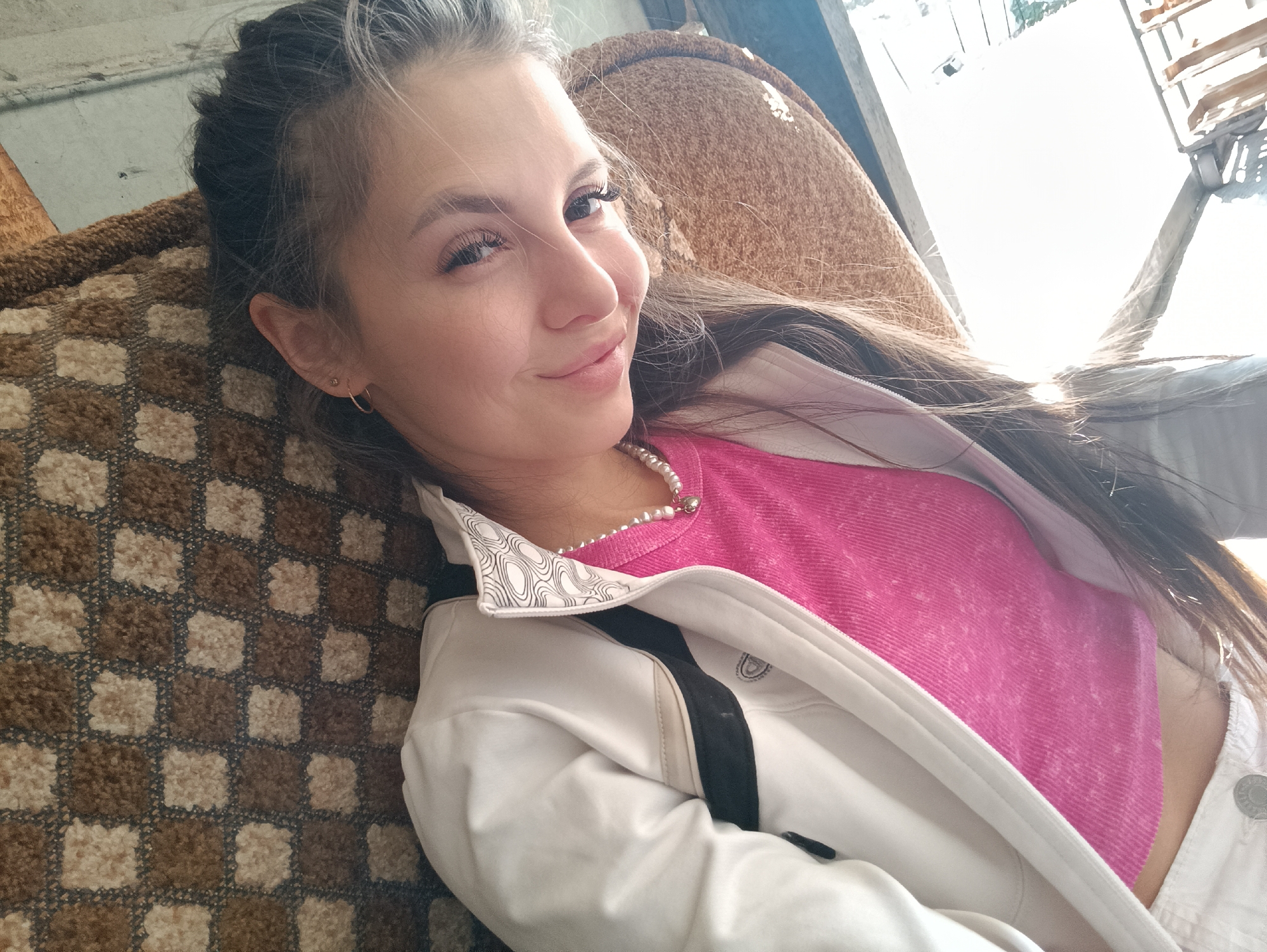 A young woman with dark hair streaked with gray and wearing a pink shirt, a white jacket, and a pearl necklace, is lying on a brown checkered upholstered surface, possibly a couch or a bed. She is smiling slightly at the camera. The background shows a blurry outdoor scene, indicating she is possibly near a window or an open space. The location in the city is unknown, unknown.