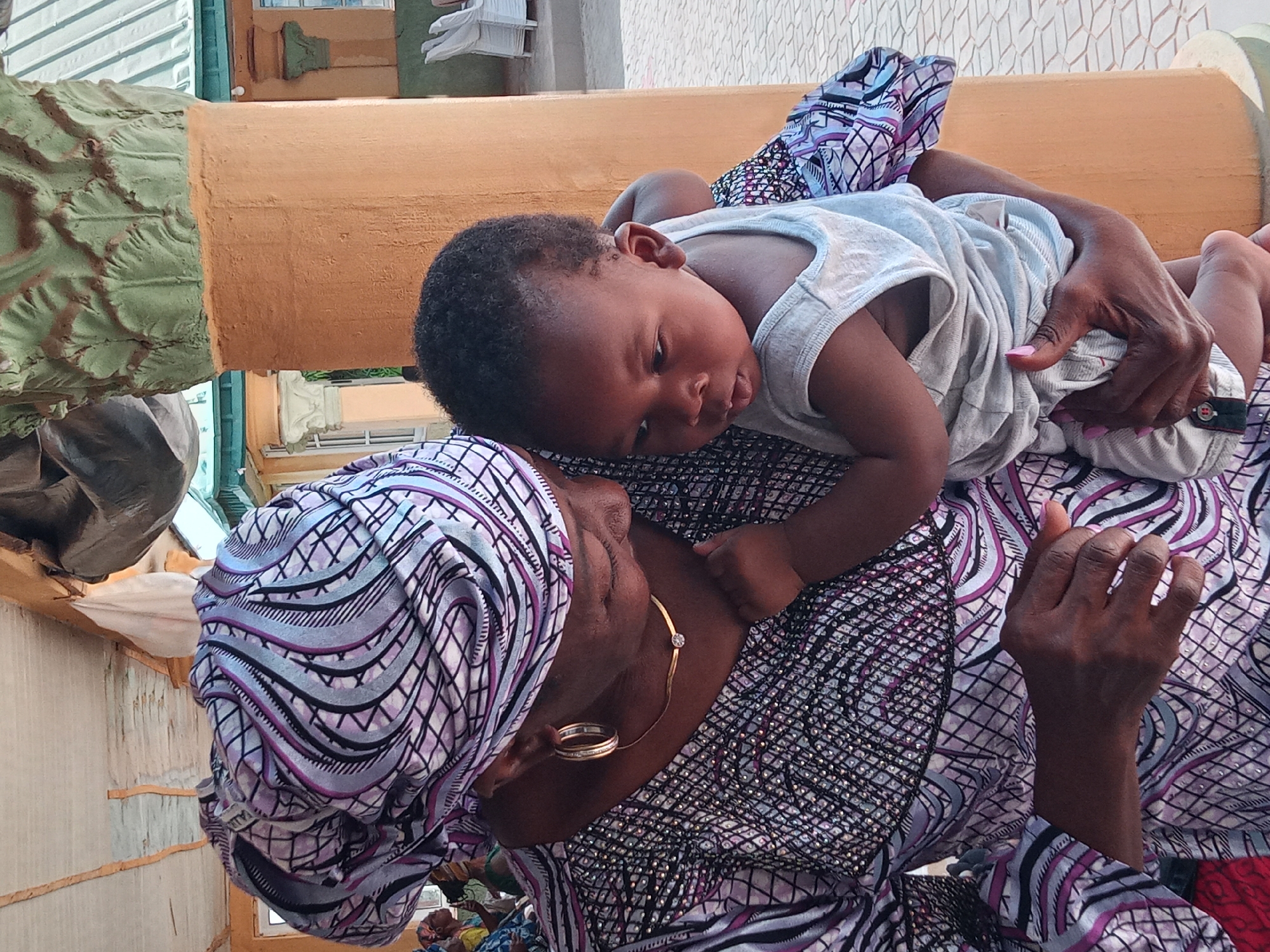 A medium shot shows a baby resting on a woman's chest, both indoors in Rido, Nigeria. The baby, dark-skinned, is lying on its side, facing left. It wears a light-colored tank top and shorts. The woman, also dark-skinned, is wearing a patterned headwrap and a patterned top in shades of white, purple, and black. A gold necklace is visible on her neck. Her hands are visible, resting on the baby. The background shows interior architectural features, including a faux-painted cylindrical support column and other elements of a building.