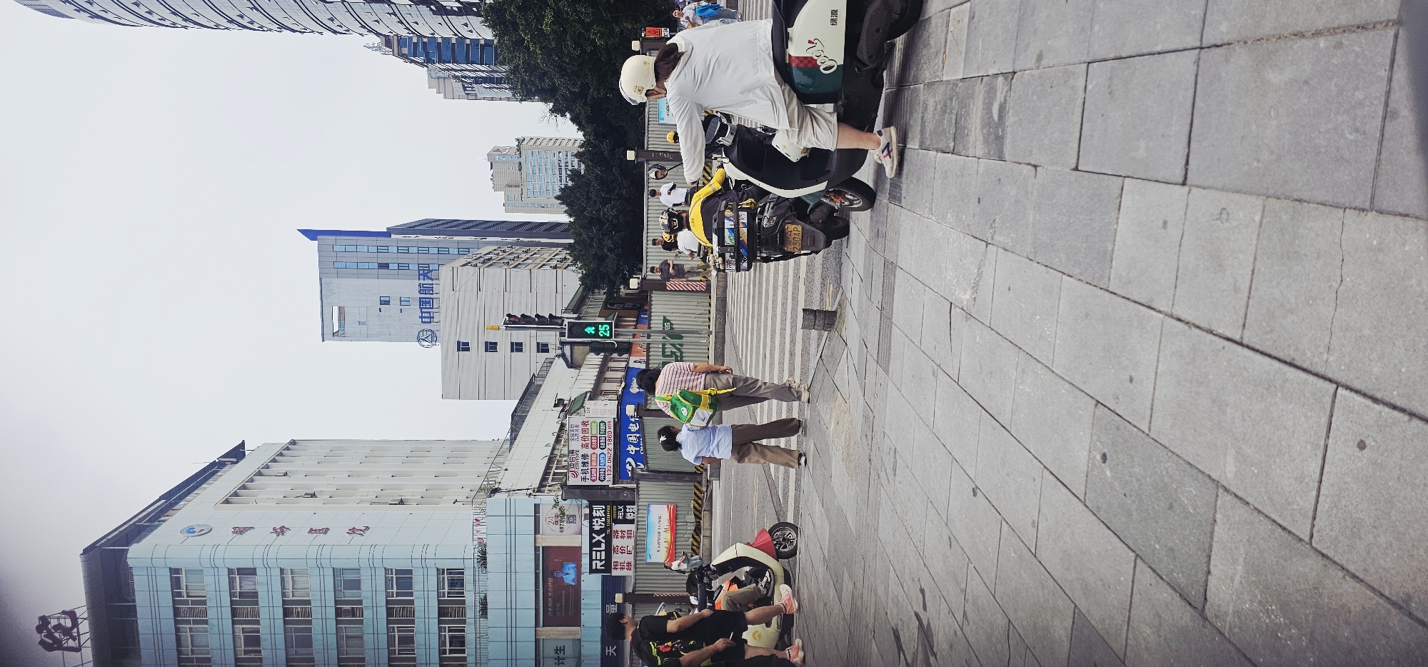 The image is a street scene in Jiulongpo District, China. Several buildings of varying heights line the street, with signs and advertisements visible on their facades. People are walking and riding scooters on the paved sidewalk and in the crosswalk. A traffic light shows a green signal. The sky is overcast.