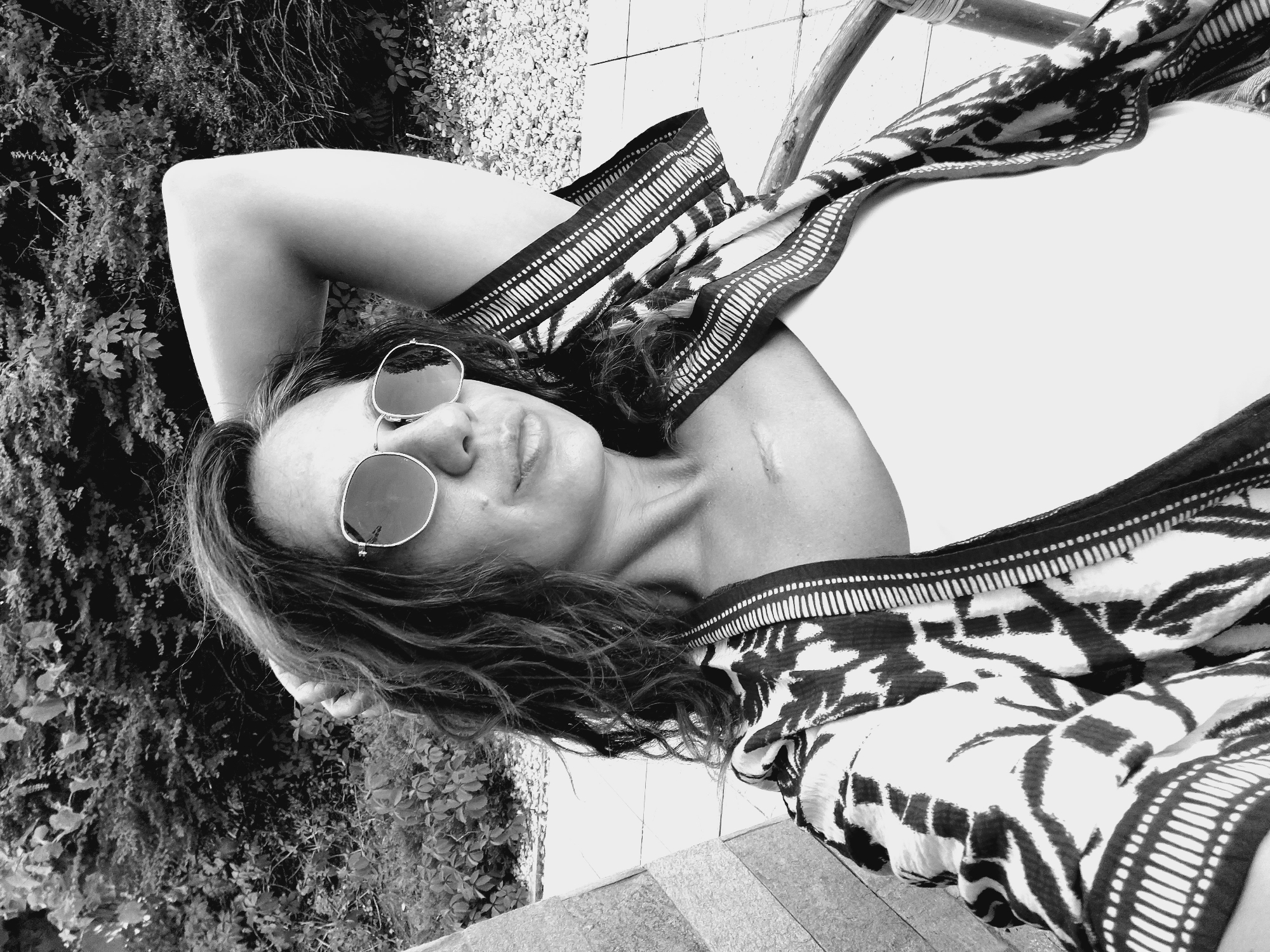A grayscale, medium shot captures a woman from the chest up, reclining outdoors. She has dark, curly hair and is wearing large, round sunglasses. Her right arm is raised and bent behind her head, with her hand resting on her hair. She is wearing a white tank top underneath a patterned kimono-style robe. A faint scar is visible on her upper chest. The background consists of lush green foliage and a stone patio, suggesting an outdoor setting. The image is likely taken in Sozopol, Bulgaria, given the mention of the city.