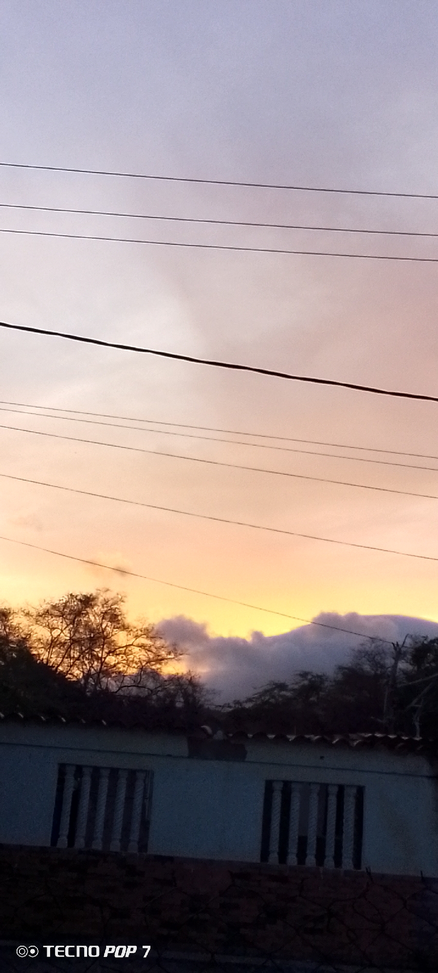 The image depicts a sunset over a building in Altagracia, Venezuela. The sky transitions from light purple at the top to orange near the horizon. Silhouetted trees and a mountain are visible against the sunset. Utility lines stretch across the sky. The building is a single story with two windows. The building has red tile roofs. A "Tecno Pop 7" brand watermark is present.
