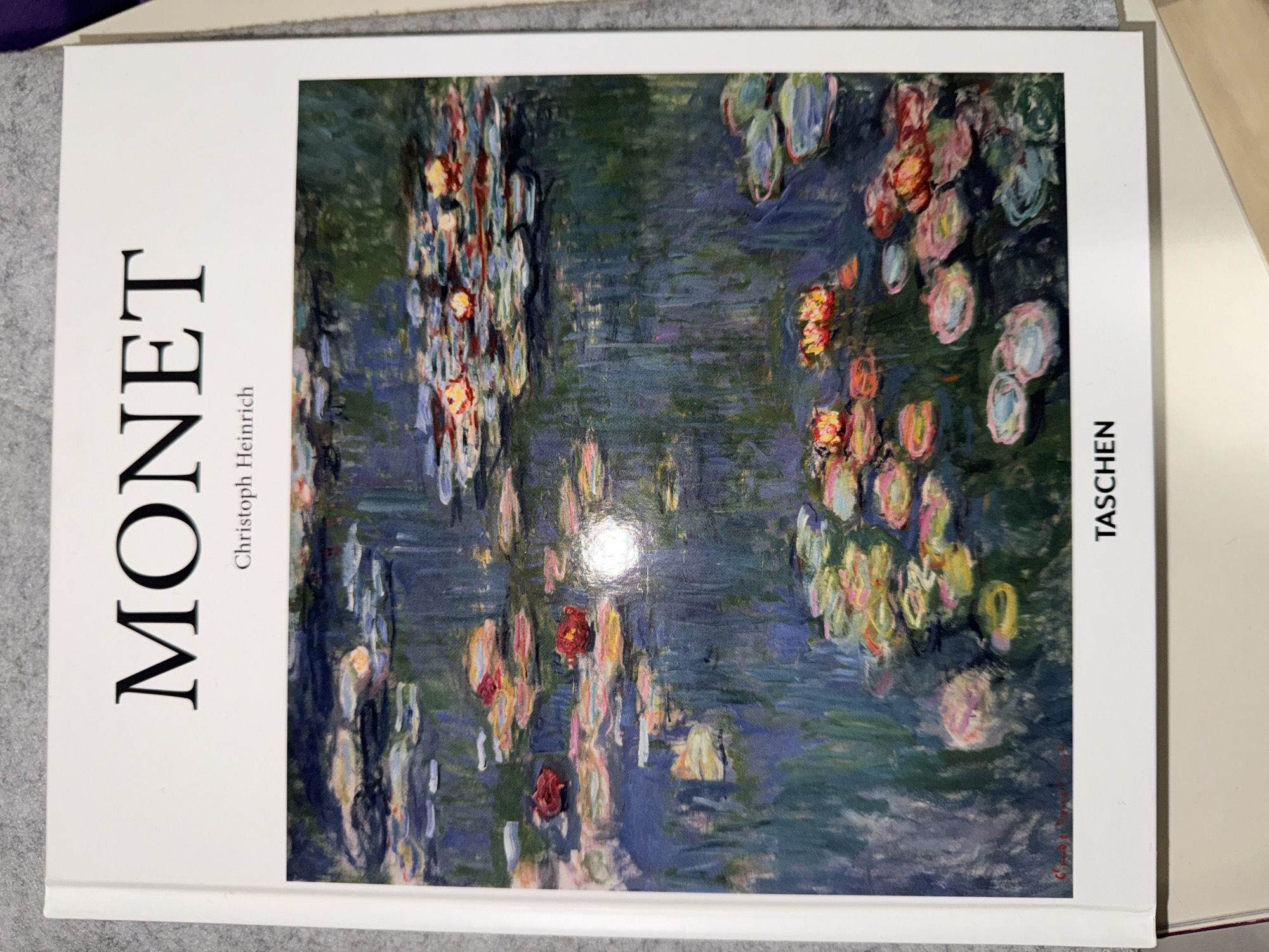 A hardcover book featuring the name "MONET" prominently on its spine-like left side and "TASCHEN" on its right edge is depicted. The book's front cover displays a vibrant, impressionistic painting, characteristic of Claude Monet's "Water Lilies" series. The artwork fills the majority of the cover, enclosed by a thin white border.
The painting features a serene water scene, dominated by shades of deep blue and green, representing the water and reflections of foliage. Scattered across the water's surface are numerous water lilies and lily pads, rendered with visible brushstrokes and a rich palette of pinks, reds, yellows, and subtle whites. Some clusters of lilies are more prominent, showcasing the interplay of light and color on the water. A noticeable glare is visible on the glossy surface of the book cover, primarily in the lower center of the artwork, reflecting ambient light.
Below the large "MONET" text, the author's name, "Christoph Heinrich," is printed in smaller black letters. The book is oriented vertically, with its long side running from the top to the bottom of the image. It rests on a light grey, textured surface, possibly fabric, with a bright white surface visible to its right. The book's crisp white edges and spine are visible, suggesting it is a new or well-preserved edition. The location of the book is Kyiv, Ukraine.