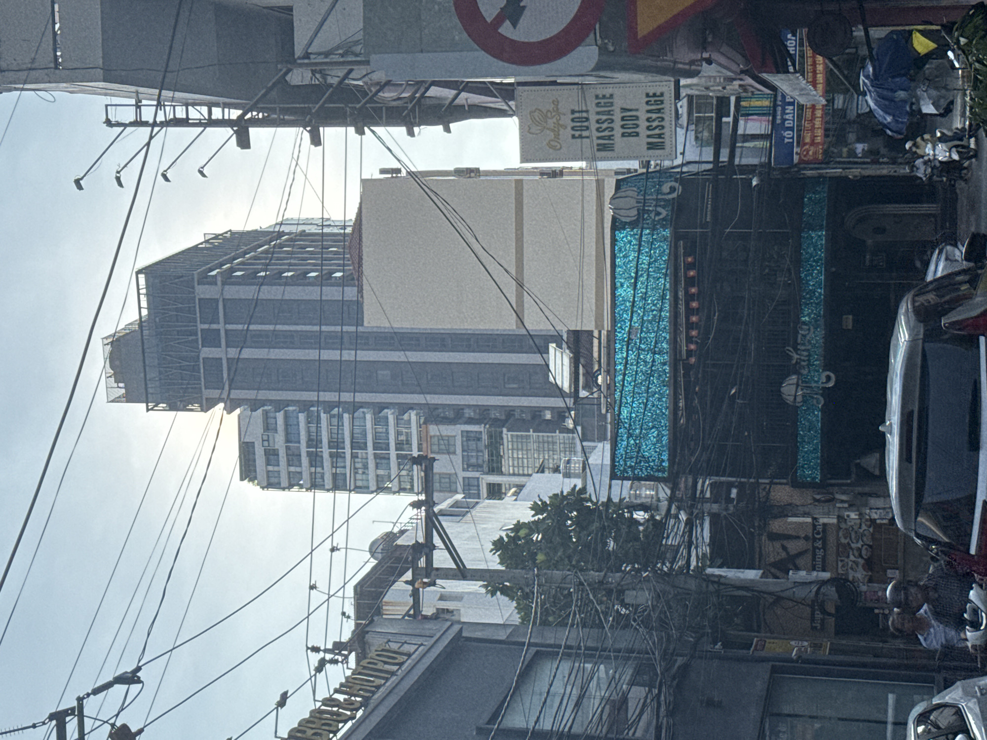 This is a vertical, eye-level, medium shot of a city street in Nha Trang, Vietnam, with a focus on buildings, signage, and electrical wires. The sky is overcast, and the lighting suggests it is daytime. On the left, a building with a sign that reads "BBQ & HOTPOT" is partially visible. The center of the image is dominated by a tall, modern building with a distinctive vertical striped facade. Below this, a darker building features bright blue, textured panels and ornate lighting fixtures. To the right, a sign for "Only Spa FOOT MASSAGE BODY MASSAGE" is prominently displayed, alongside a red and white circular sign with an arrow indicating a turn. Various other signs, some in Vietnamese, are visible on the buildings. Numerous electrical wires crisscross the scene, indicating an urban environment with extensive utility infrastructure. In the foreground, two individuals on a motorbike are partially visible, along with the roof of a car. The overall impression is one of a busy, somewhat cluttered urban streetscape typical of Southeast Asia.