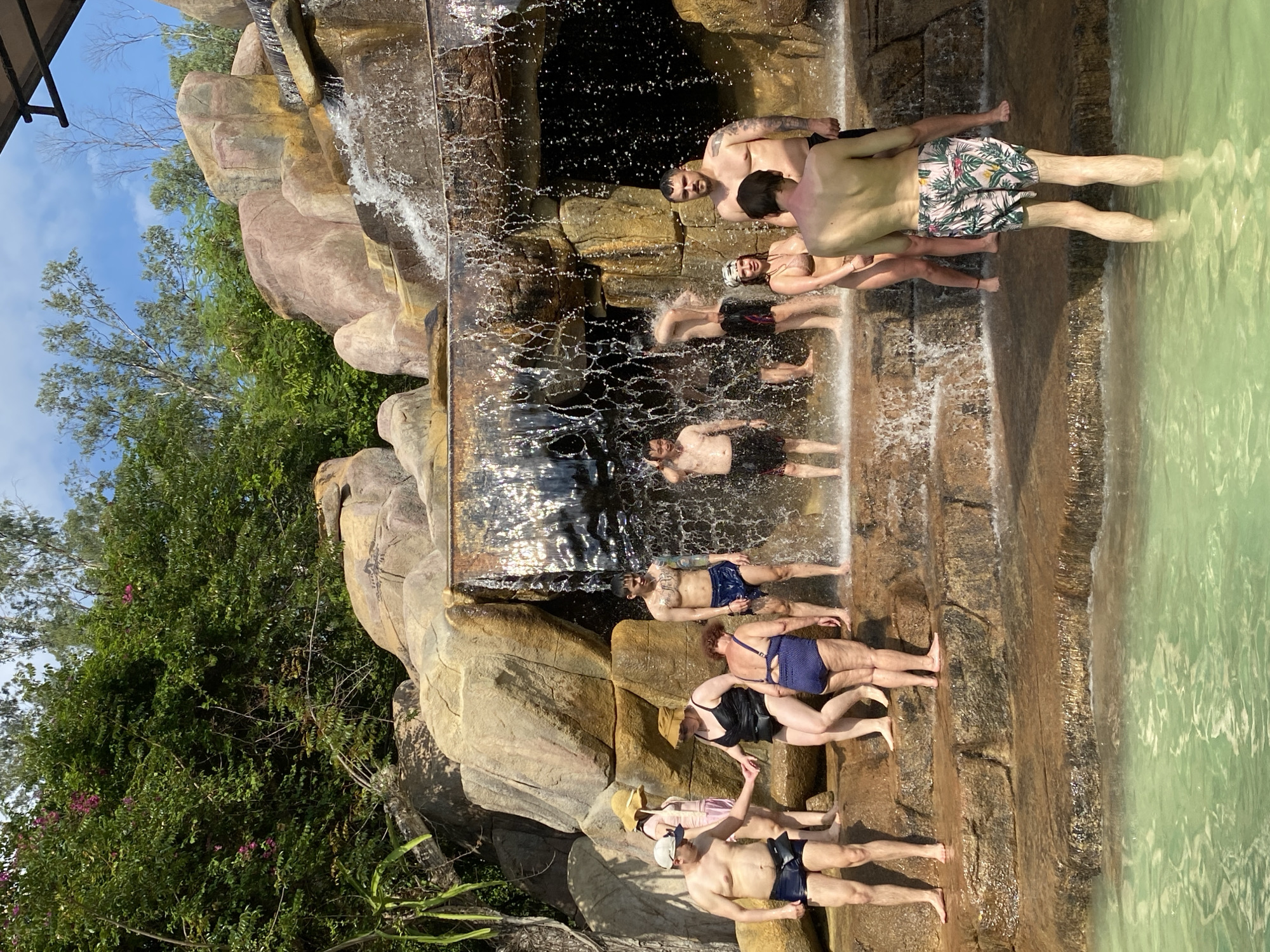 In the city of Nha Trang, Vietnam, a group of people are enjoying themselves at a water park. The scene unfolds at the base of a man-made waterfall, composed of large, rough-hewn rocks. The water cascades down, creating a refreshing mist. Several individuals are standing on the tiered, rocky platforms surrounding the waterfall, while others are partially submerged in the turquoise water. The people are dressed in swimwear, suggesting a leisurely day of recreation. Lush green foliage borders the top of the waterfall, adding to the tropical ambiance. The lighting indicates it is daytime, with sunlight casting shadows and highlighting the water's movement. The overall impression is one of a vibrant and lively tourist destination.
