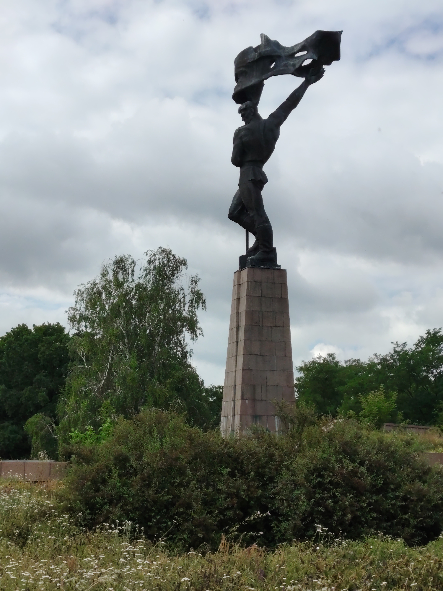 The image shows a statue of a man holding a flag against a cloudy sky. The statue is made of dark metal and stands on a tall, stone pedestal. The man is depicted in a dynamic pose, with his arm raised, holding a tattered flag that appears to be blowing in the wind. The statue is located in the village of Balyko-Shchuchynka, Ukraine, a site of historical significance related to the Second World War. The foreground of the image is filled with dense green bushes and scattered white wildflowers. In the background, several trees with green foliage are visible, along with a portion of a concrete barrier. The overall scene is somber, with the overcast sky contributing to a dramatic atmosphere.