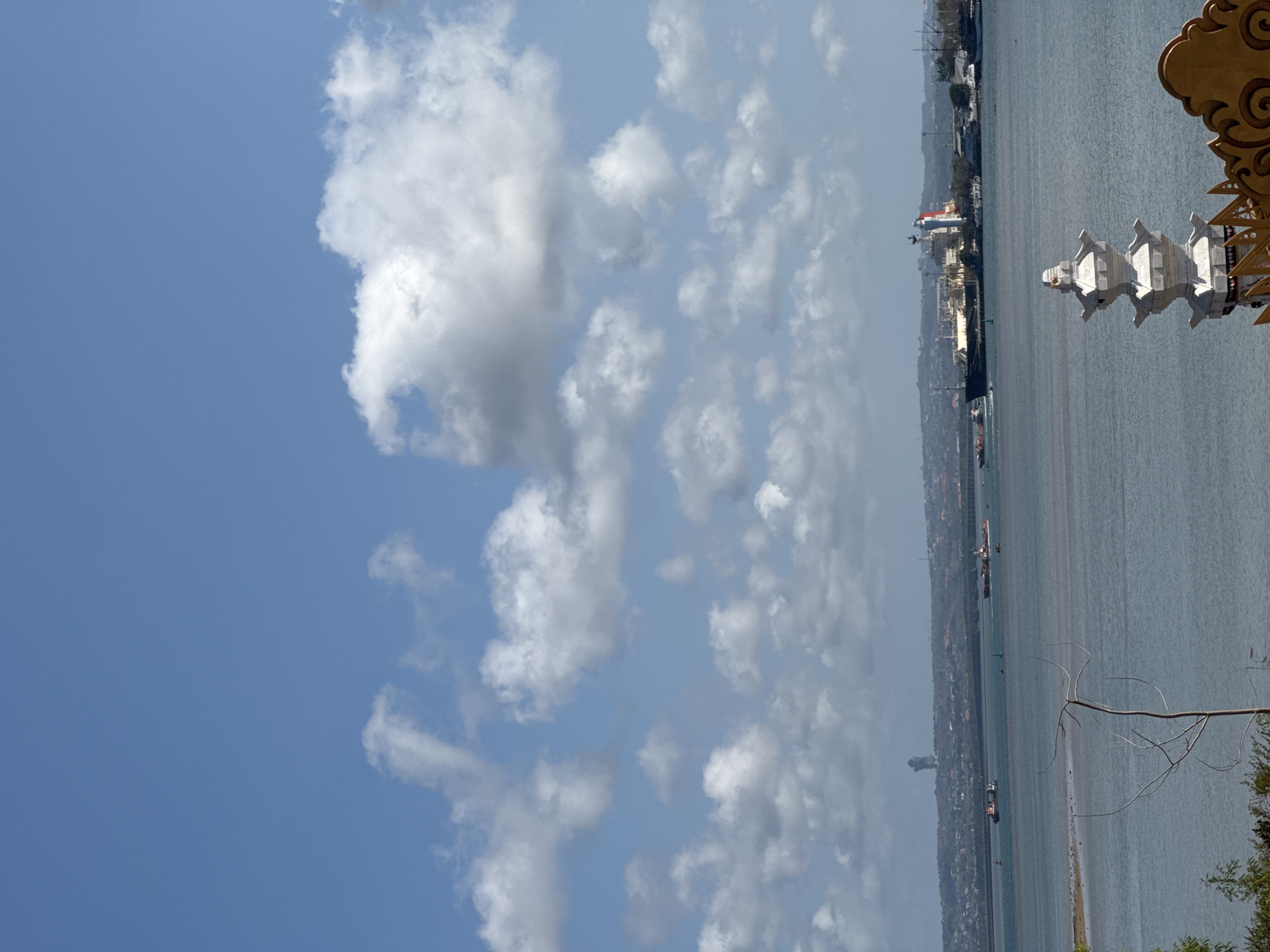 A wide-angle, eye-level outdoor shot captures a serene seascape under a partly cloudy sky, likely depicting a harbor or coastal area in Indonesia, given the architectural style of the foreground structures and the presence of large ships. The sky is a vibrant blue, dotted with fluffy white cumulus clouds, some of which are larger and more defined than others.
The middle ground of the image features a calm, light blue body of water, reflecting the sky and clouds. Several ships are visible on the water: on the left, two smaller vessels appear to be moving. Further to the right, a large cargo ship, likely a tanker, is docked or anchored near a pier, identifiable by its white and red structure. Other, smaller boats and buoys are also scattered across the water.
The background presents a hazy, distant shoreline lined with buildings and greenery, suggesting an urban or semi-urban coastal settlement. A tall, dark, irregular-shaped structure, possibly a tower or antenna, rises prominently against the skyline in the far distance.
In the foreground, a white, multi-tiered architectural element, resembling a stupa or pagoda, is positioned on the right. Its intricate, stacked design is a focal point, with ornate golden detailing visible on the far right edge of the frame. The lower left foreground shows a bare tree branch extending into the frame from the left, adding a touch of natural detail against the man-made and aquatic elements. The overall impression is one of a calm, sunny day at a bustling port or coastal town.