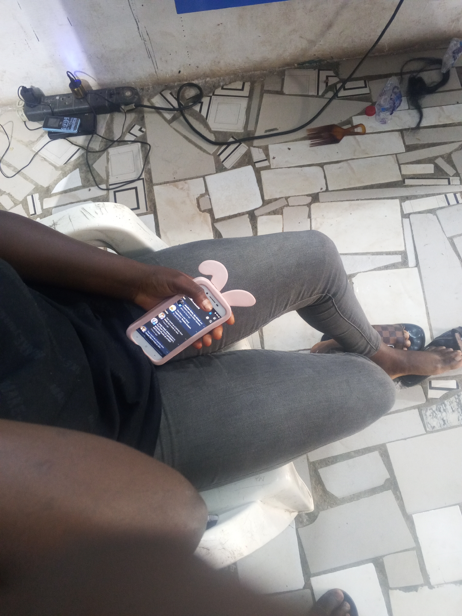 A person is seated in a white plastic chair, holding a pink smartphone with bunny ear decorations on its case. The screen displays what appears to be social media notifications. The person is wearing grey jeans and black sandals. The floor is tiled with irregularly shaped white tiles. In the background, there is a power strip with connected plugs, a water bottle, and a hair pick. The location is determined to be Gwong, Nigeria based on the presence of "PLATEAU STATE UNIVERSITY" and other information visible on the phone screen.