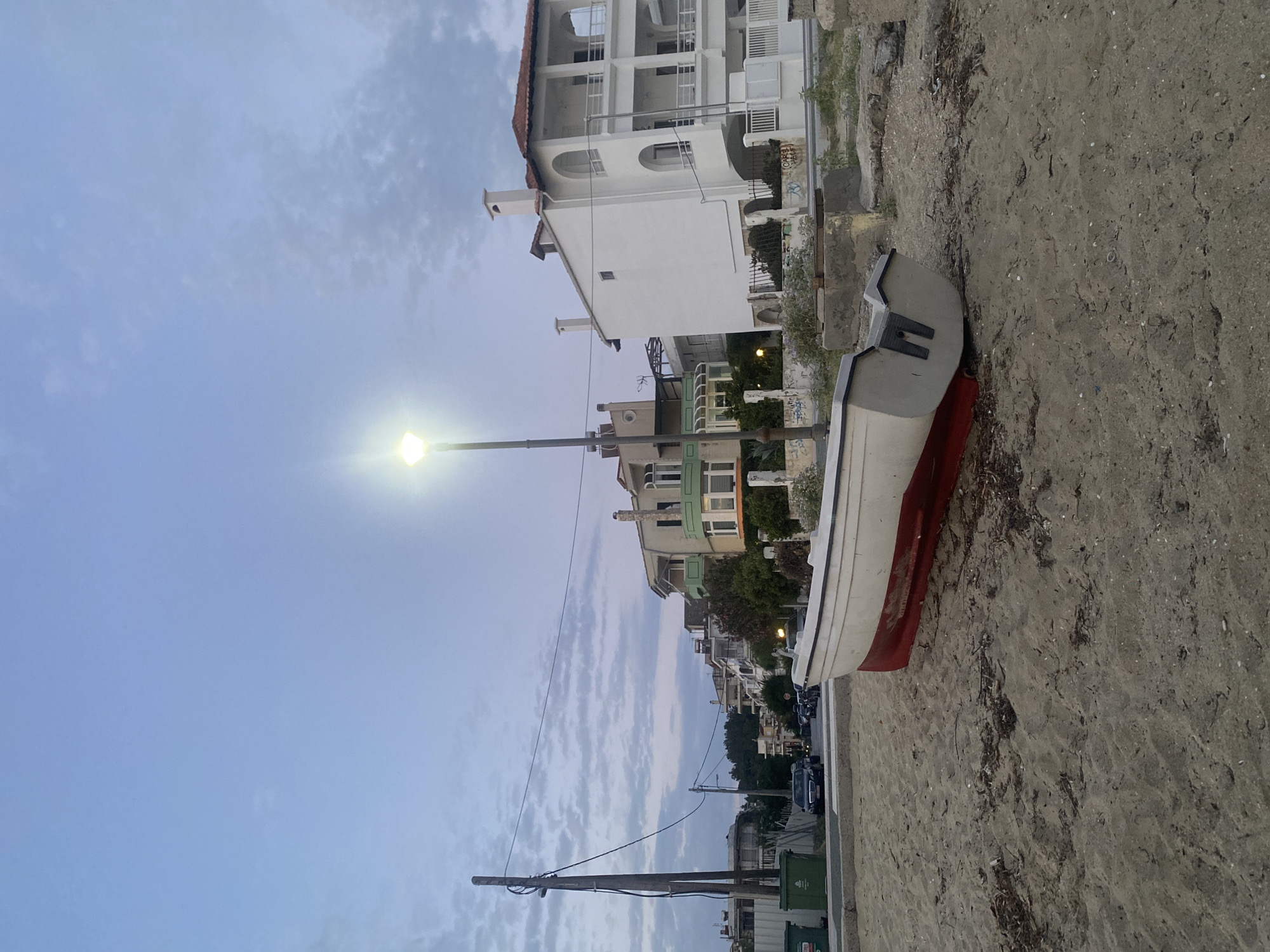 The image depicts a beach scene in Neoi Epivates, Greece, at dusk. In the foreground, a white boat with a red hull rests on the sandy beach. The boat is positioned at an angle, with its stern slightly elevated. The sand is mixed with small debris.
In the midground, a street runs parallel to the beach, lined with several multi-story buildings. These buildings feature balconies, arched windows, and red-tiled roofs, characteristic of Mediterranean architecture. A street lamp illuminates the scene, casting a bright light downwards. There are power lines and utility poles visible along the street. Cars are parked and driving on the street. Two green trash bins are also visible on the left side of the street.
The background shows more buildings and trees, extending into the distance. The sky is a soft blue with scattered clouds, indicating the transition from day to night. The overall atmosphere is calm and serene.
